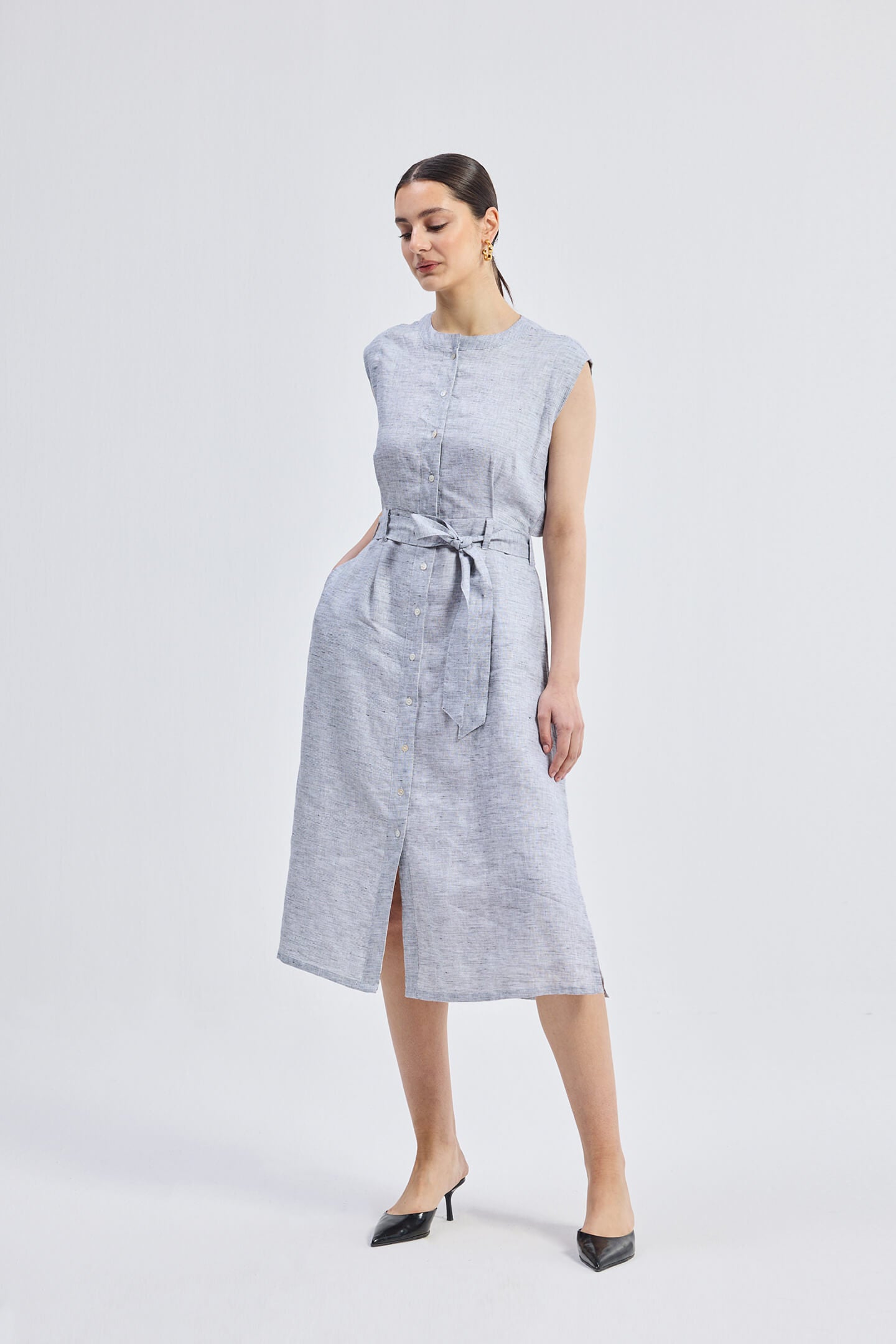 Full-length front view of model wearing the Reistor Back Cut Out Linen Dress, featuring a sleeveless design, button-down front, tie-waist belt, and light blue striped linen fabric.