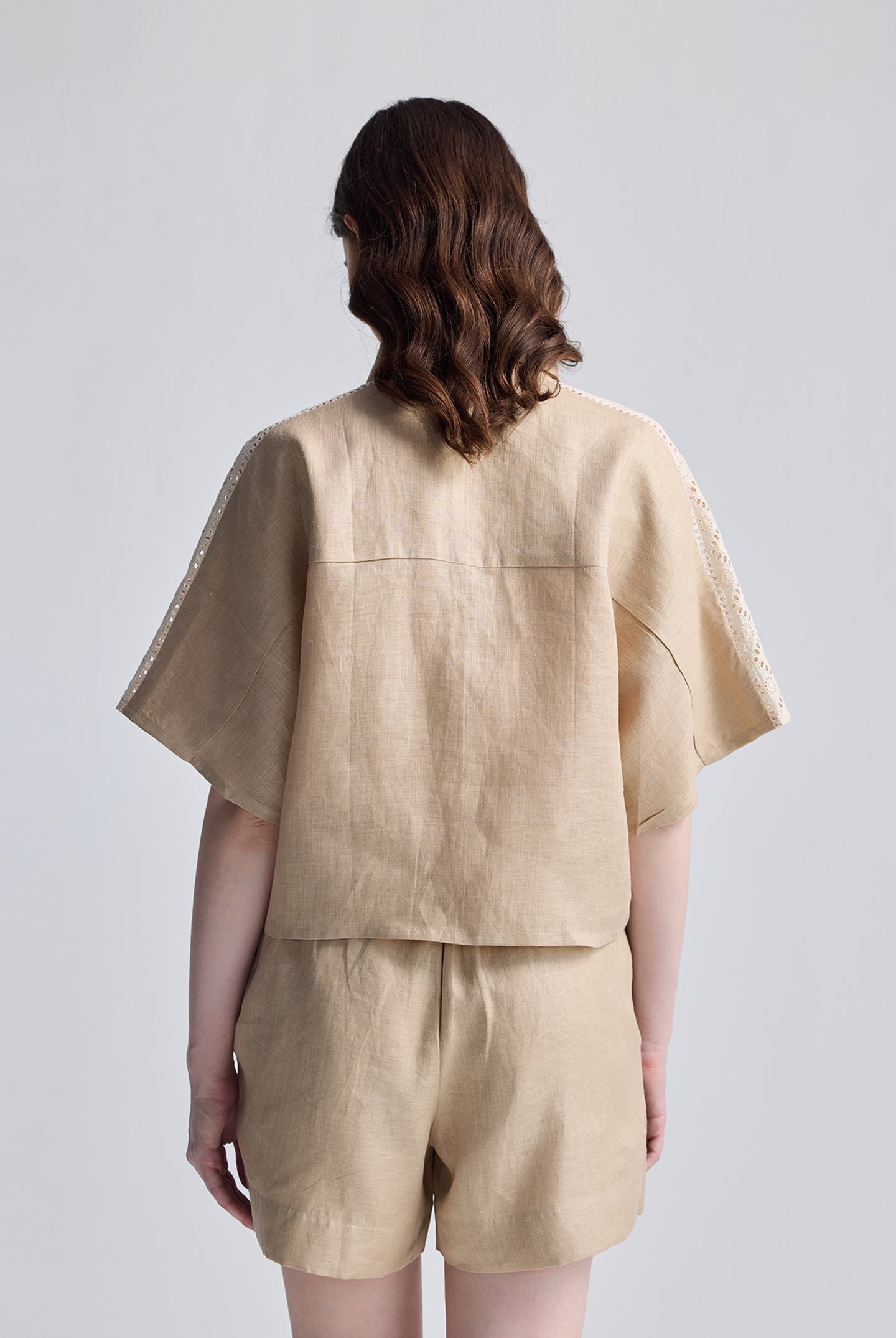 Back view of a model wearing the Boxy Shirt with Lace Set by reistor, showing a beige hemp-blend short-sleeve shirt with delicate white lace detailing on the shoulders and matching relaxed shorts.