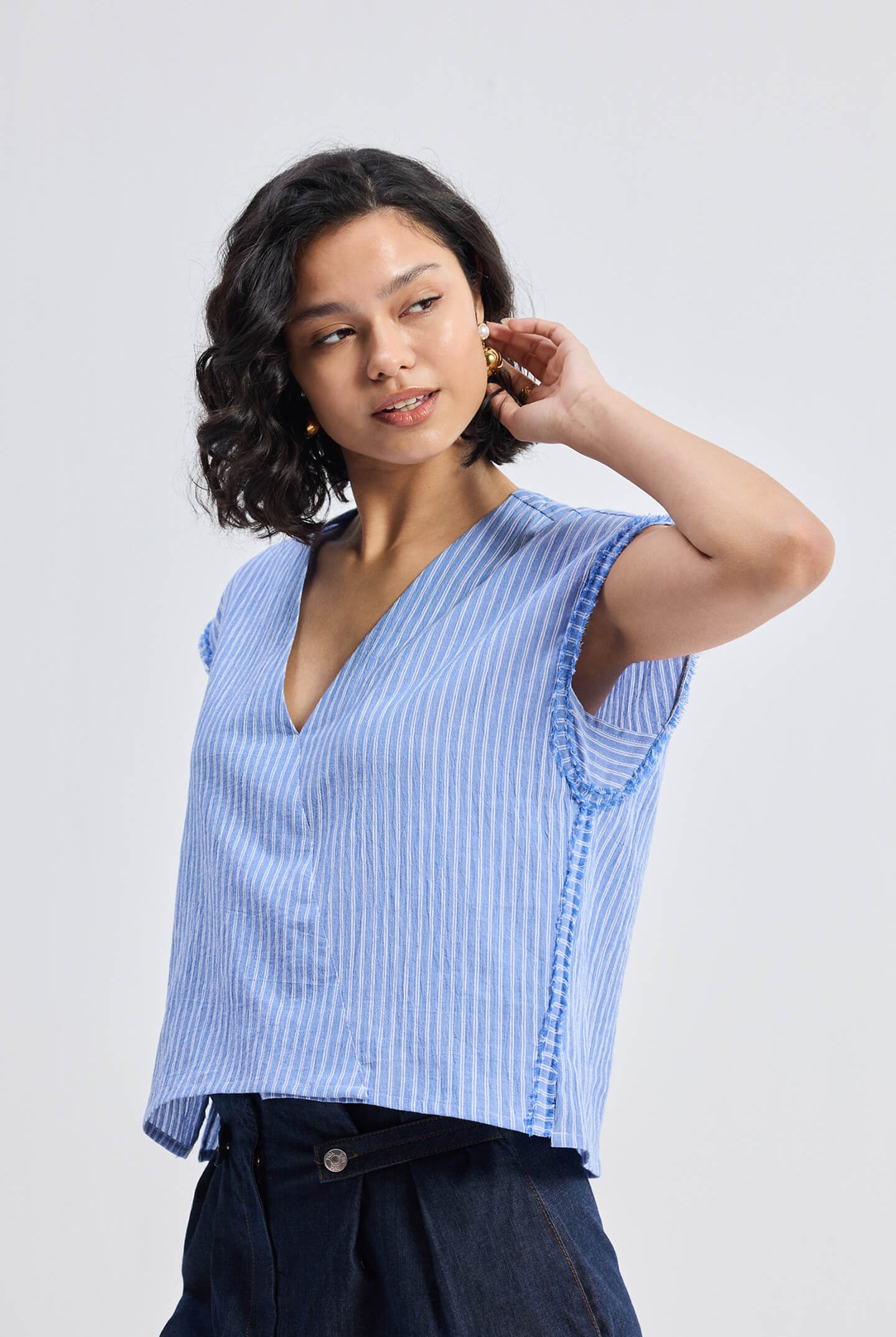 Side view of model wearing Reistor Boxy V-neck Top with Fray Detail in blue striped textured fabric, relaxed fit sustainable women’s top paired with dark cuffed shorts.