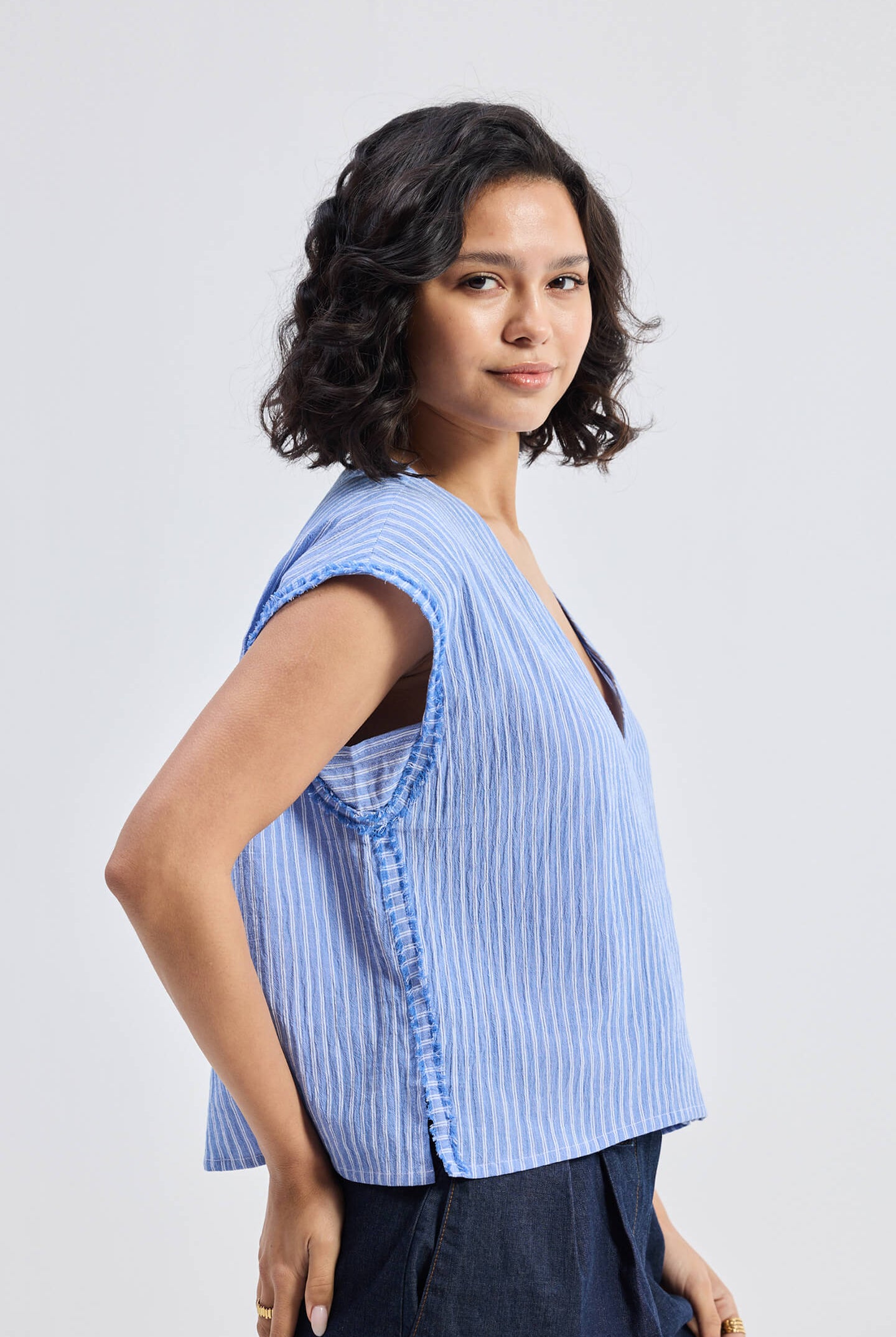 Model wearing Reistor Boxy V-neck Top with Fray Detail in blue textured fabric, relaxed fit sleeveless sustainable women’s top, side view.