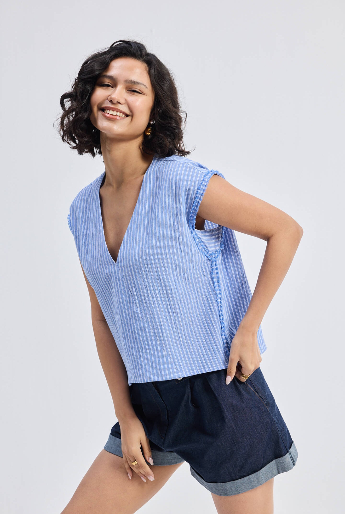 Smiling model wearing Reistor Boxy V-neck Top with Fray Detail in blue textured fabric, relaxed fit sleeveless eco-friendly women’s top styled with dark cuffed shorts.
