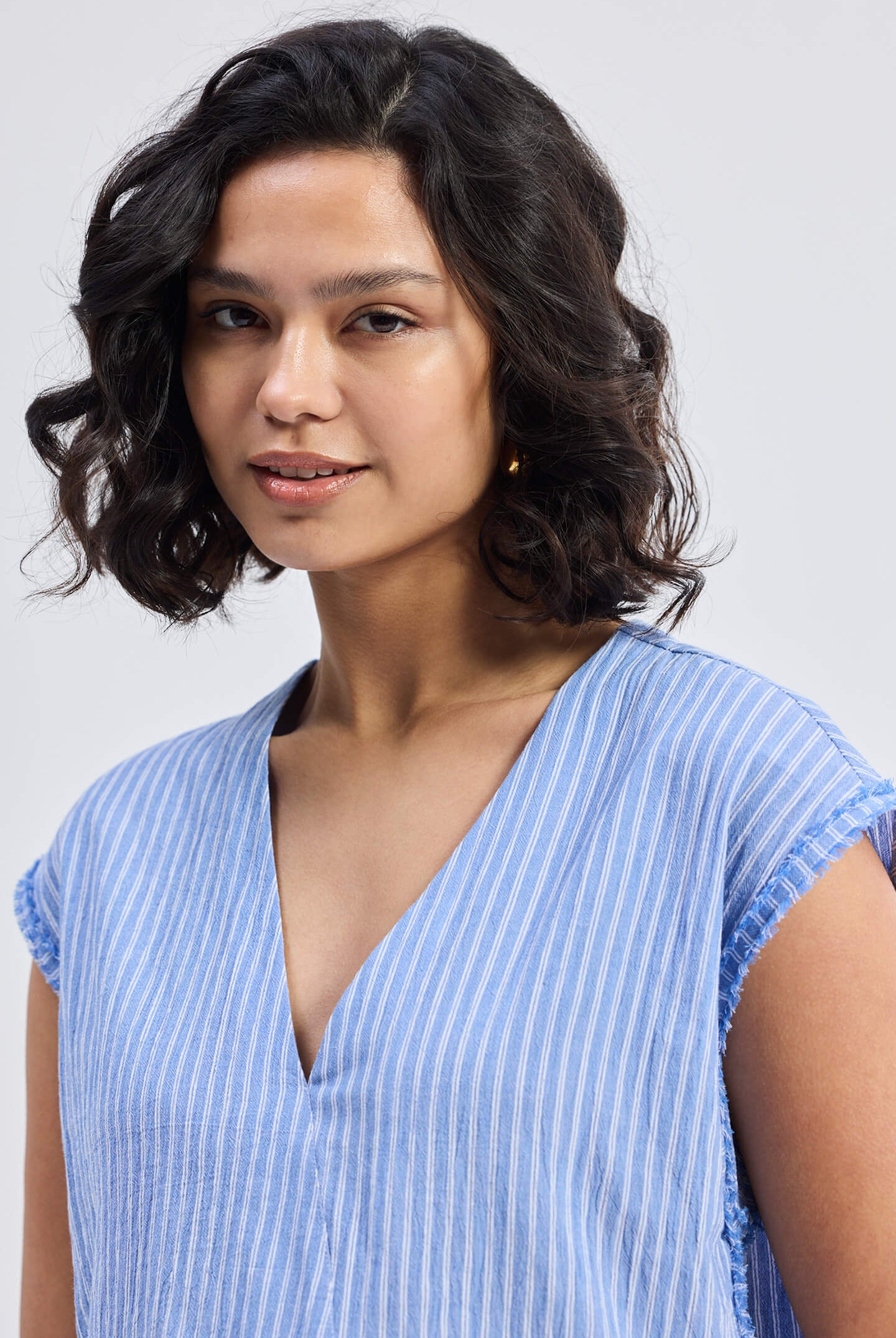 Close-up of model wearing Reistor Boxy V-neck Top with Fray Detail in blue striped textured fabric, relaxed fit sustainable women’s top with frayed sleeve edges.