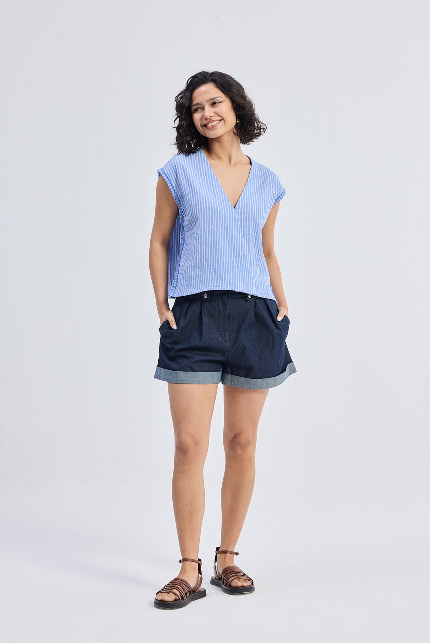 Front full-length view of model wearing Reistor Boxy V-neck Top with Fray Detail in blue striped textured fabric, relaxed fit sustainable women’s top styled with dark cuffed shorts and sandals.