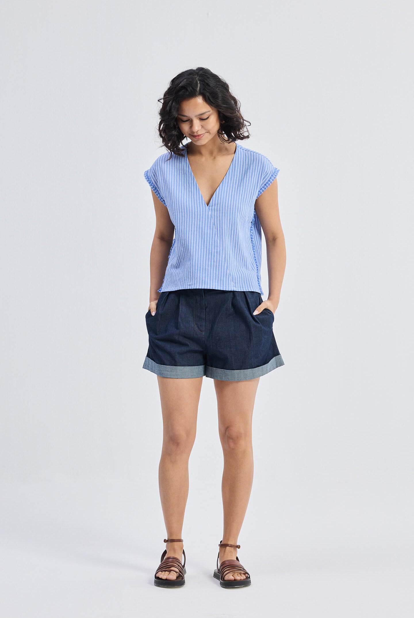 Model wearing Reistor Boxy V-neck Top with Fray Detail in blue striped textured fabric, relaxed fit eco-friendly women’s top paired with dark cuffed shorts and sandals, front view.