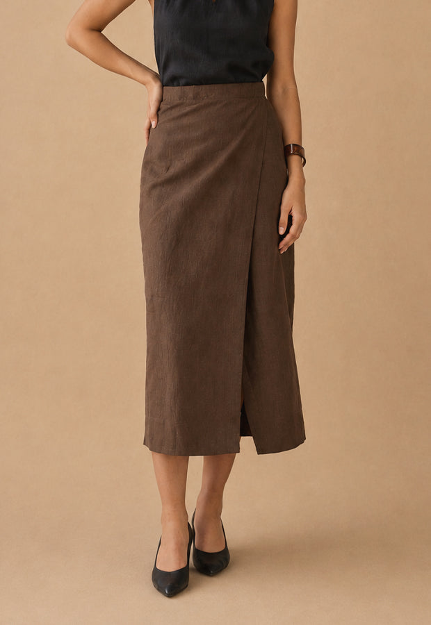 A woman models a brown wrap midi skirt with a side slit, paired with a black sleeveless top and black heels, against a solid beige studio background.