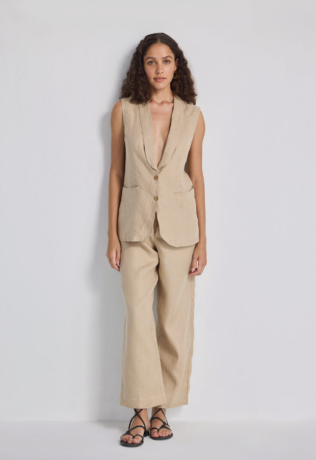 A woman modeling a beige linen two-piece suit consisting of a sleeveless V-neck blazer and matching wide-leg trousers, paired with black strappy sandals, against a light gray studio background.