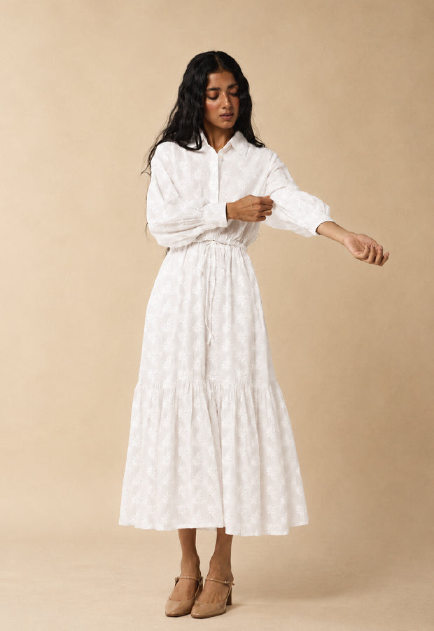A woman modeling a white long-sleeve midi dress with a subtle tonal pattern, featuring a button-up collar, gathered waist, and tiered skirt, set against a solid beige studio background.