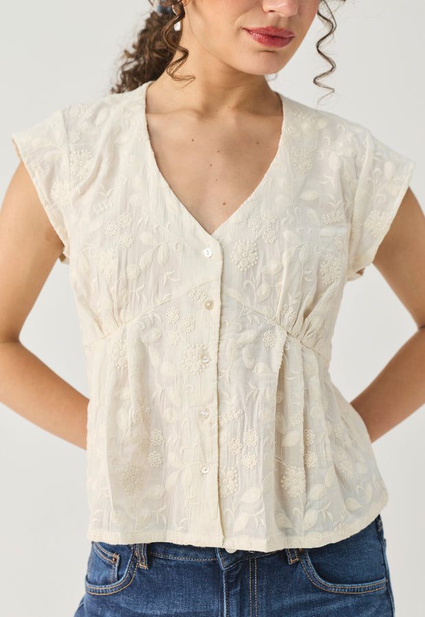 A woman wearing a cream-colored sleeveless blouse with a V-neckline, button-up front, and textured floral embroidery, paired with blue jeans against a light gray background.