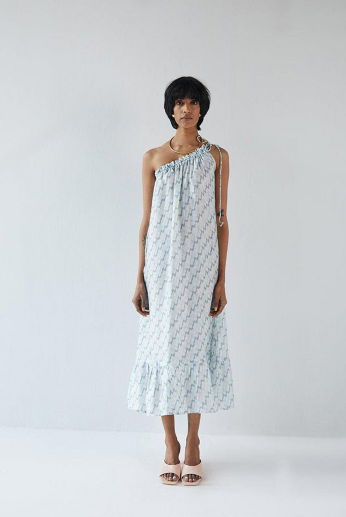 Woman wearing the Reistor Dancing in the Moonlight Dress, a light blue patterned one-shoulder sustainable dress, perfect for summer vacations, brunches, and resort wear.