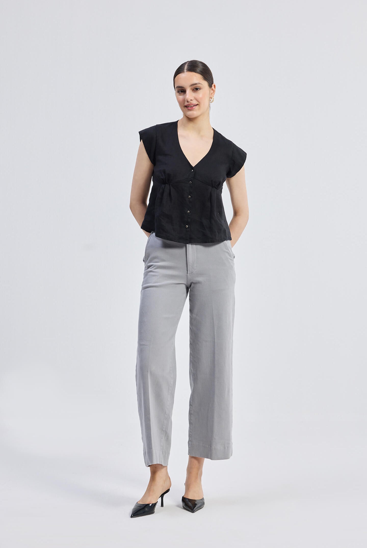 Deep V-neck short top in Black by reistor featuring cap sleeves, front button detailing, and a relaxed fit silhouette styled with grey wide-leg trousers, full-length view.