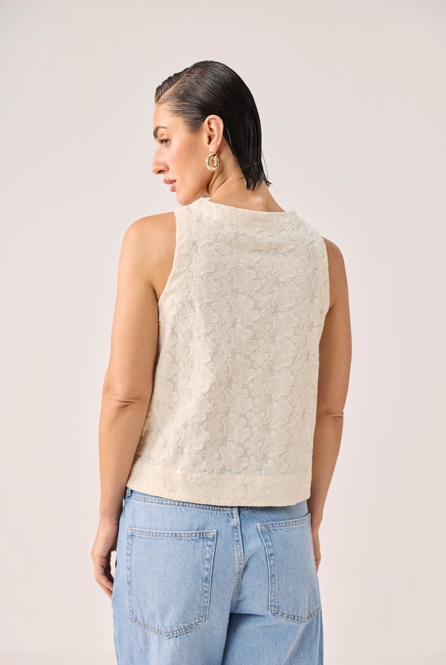 Embroidered Button Front Neck Top in Cream featuring an all-over floral embroidered texture, sleeveless silhouette, and button-front design, shown from the back and paired with light-wash denim jeans in a clean studio setting.
