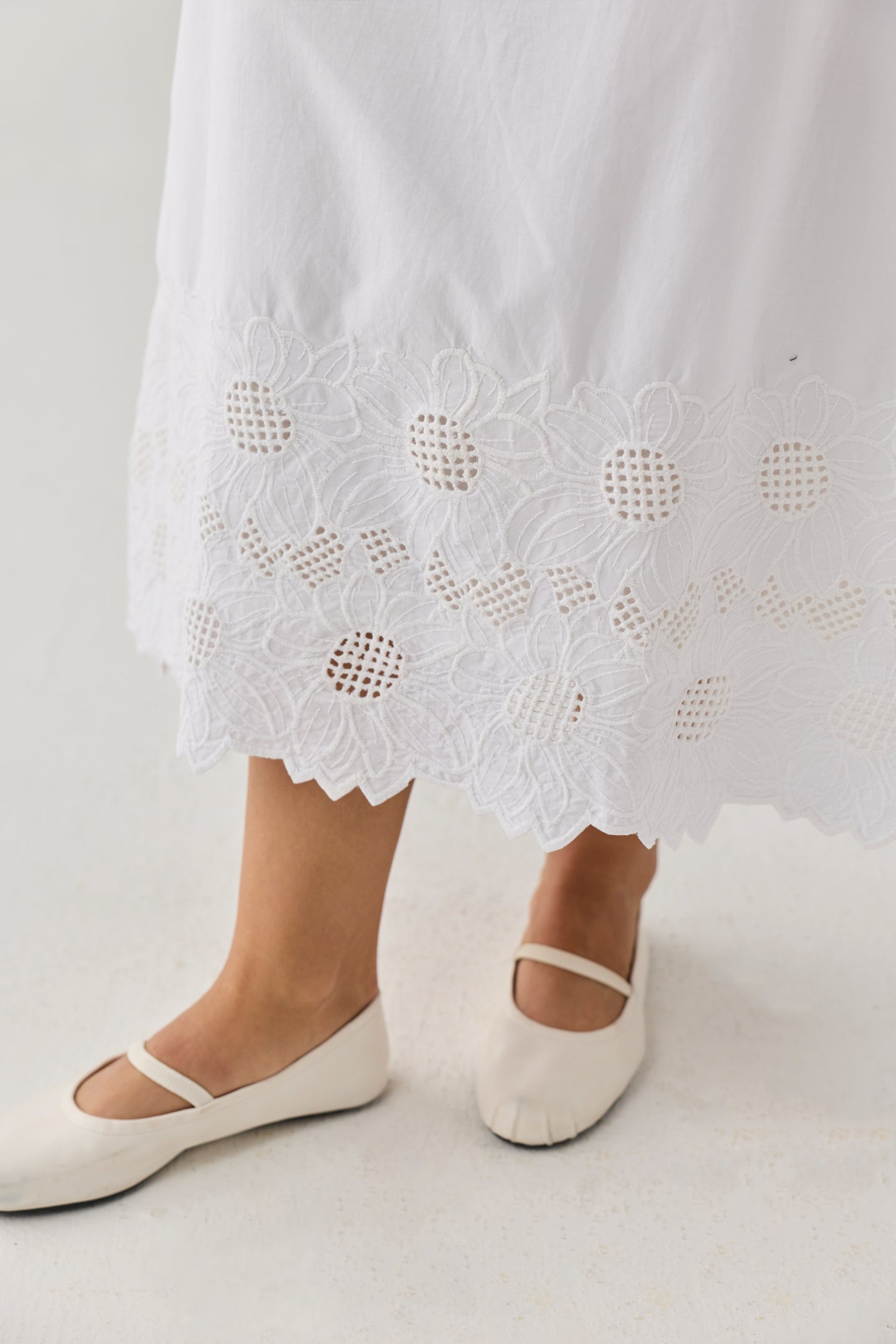 Embroidered_Midi_Skirt_in_White — White midi skirt featuring intricate floral embroidery and eyelet lace detailing along the hem, shown with white Mary Jane-style flats against a neutral background.
