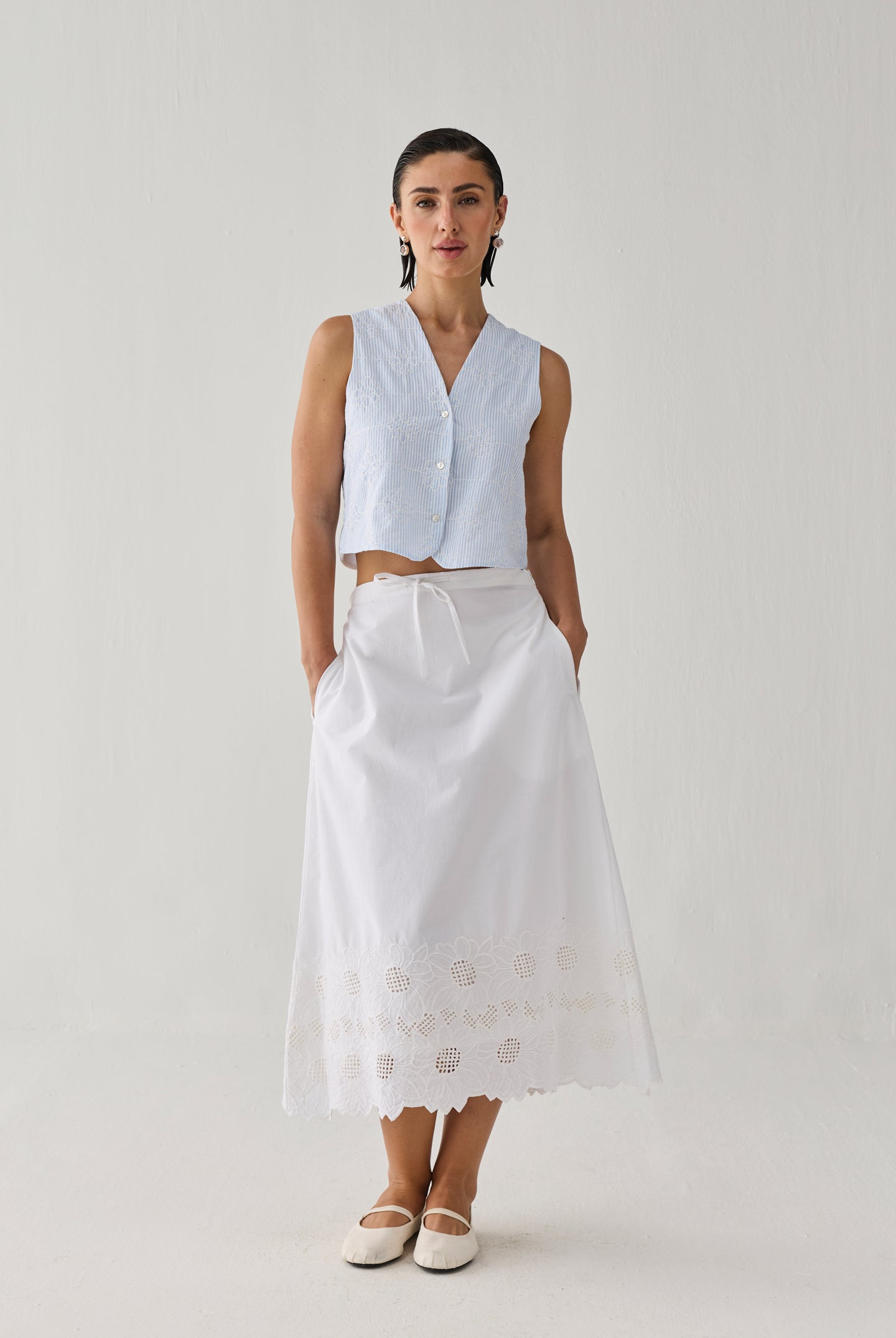 Woman wearing an embroidered white midi skirt with delicate cutwork detailing along the hem, paired with a sleeveless light blue striped top and white flats, standing against a plain light background.