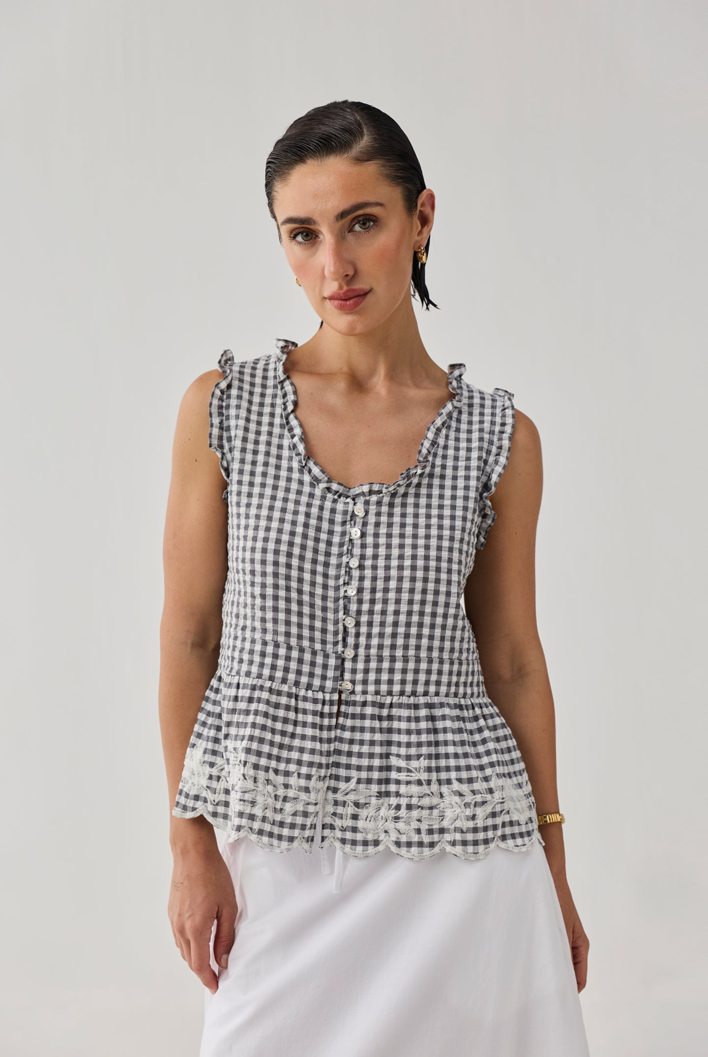 Woman wearing an embroidered peplum top in grey gingham check with ruffled trim, front button detailing, and lace embroidery at the hem, paired with a white skirt against a neutral background.
