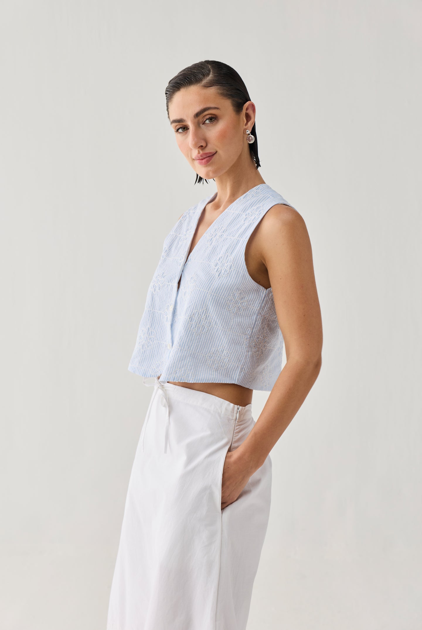 Embroidered Sleeveless Vest Top in Blue Stripe with a textured striped pattern, V-neckline, and cropped sleeveless silhouette, shown from a side angle and paired with a white skirt in a minimalist studio setting.