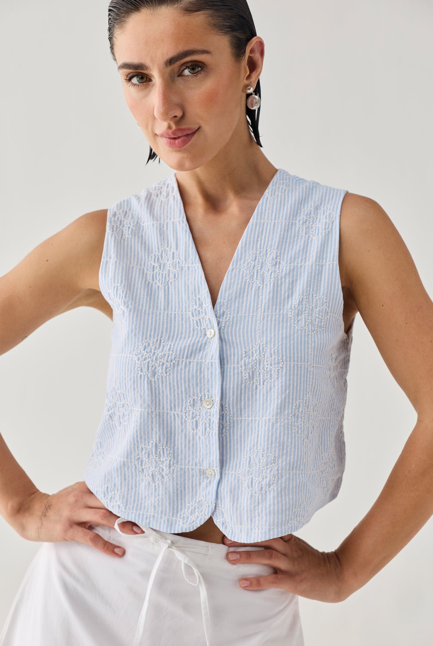Embroidered Sleeveless Vest Top in Blue Stripe featuring delicate floral embroidery, a textured striped pattern, V-neckline, and button-front design, shown in a close-up view and paired with a white skirt.