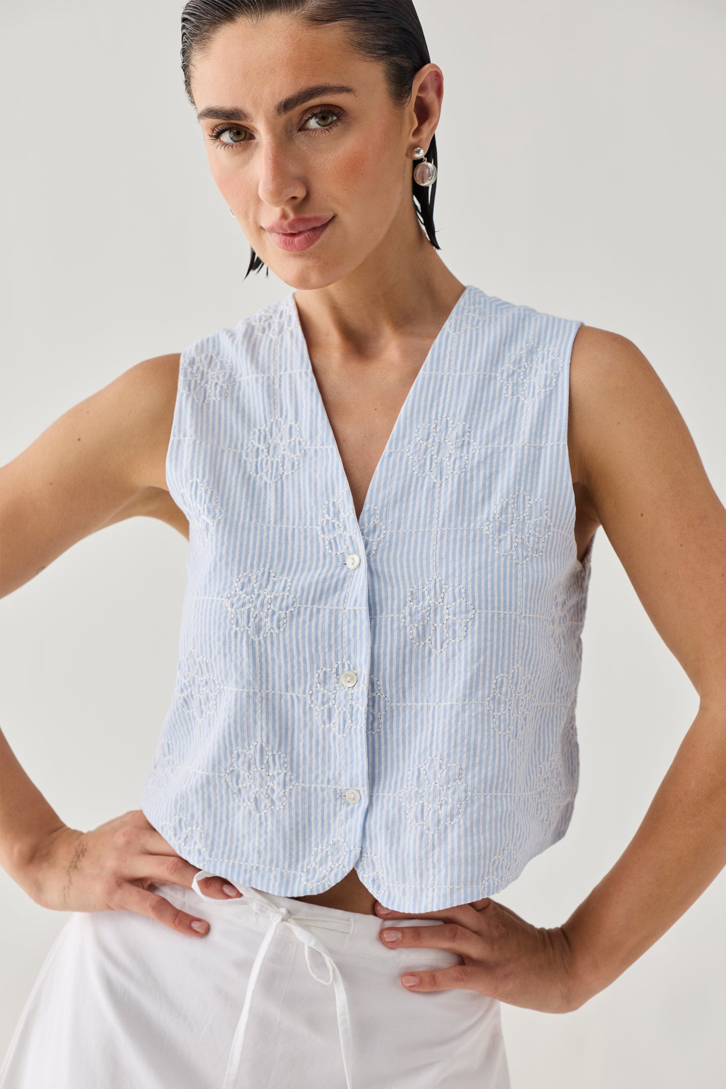 Embroidered Sleeveless Vest Top in Blue Stripe featuring delicate floral embroidery, a textured striped pattern, V-neckline, and button-front design, shown in a close-up view and paired with a white skirt.