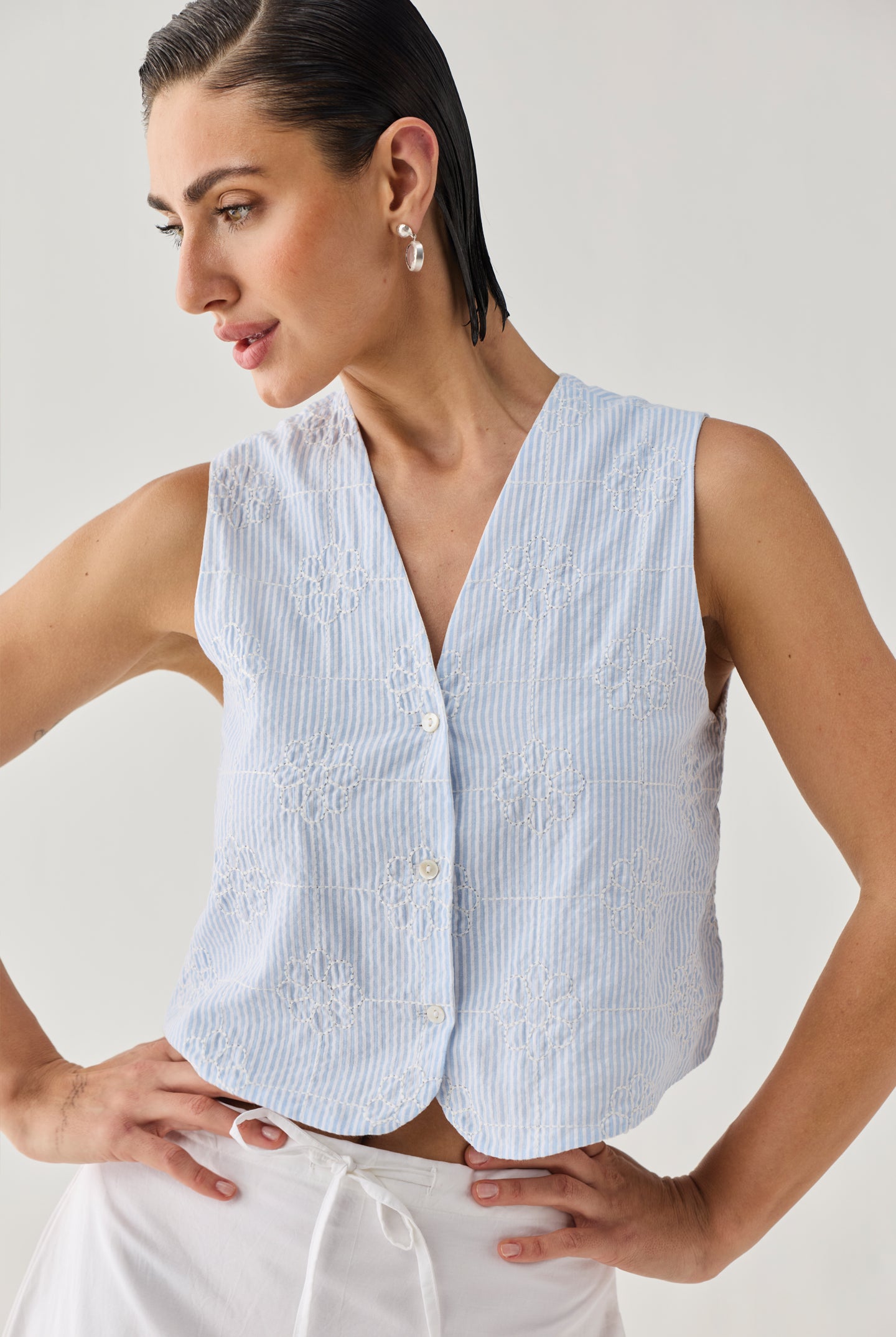 Embroidered Sleeveless Vest Top in Blue Stripe featuring delicate tonal floral embroidery, a textured striped pattern, V-neckline, and button-front design, shown from an angled view to highlight the lightweight fabric and relaxed cropped silhouette.