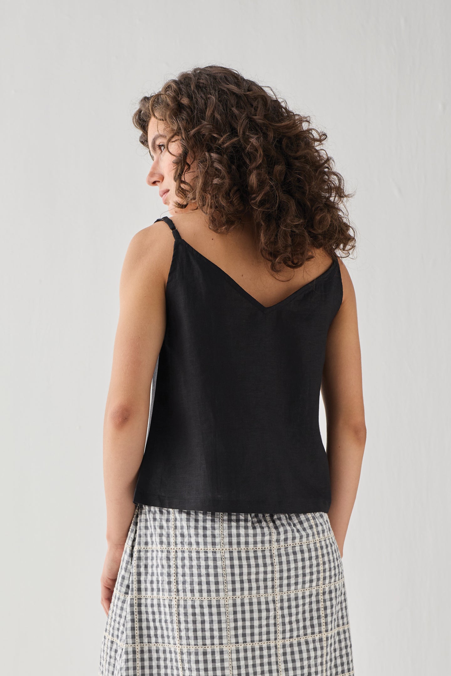 Hemp Front Tie Cami Top in Black, shown from the back, featuring a relaxed fit, thin shoulder straps, and a lightweight breathable fabric. The model pairs it with a check-pattern skirt.