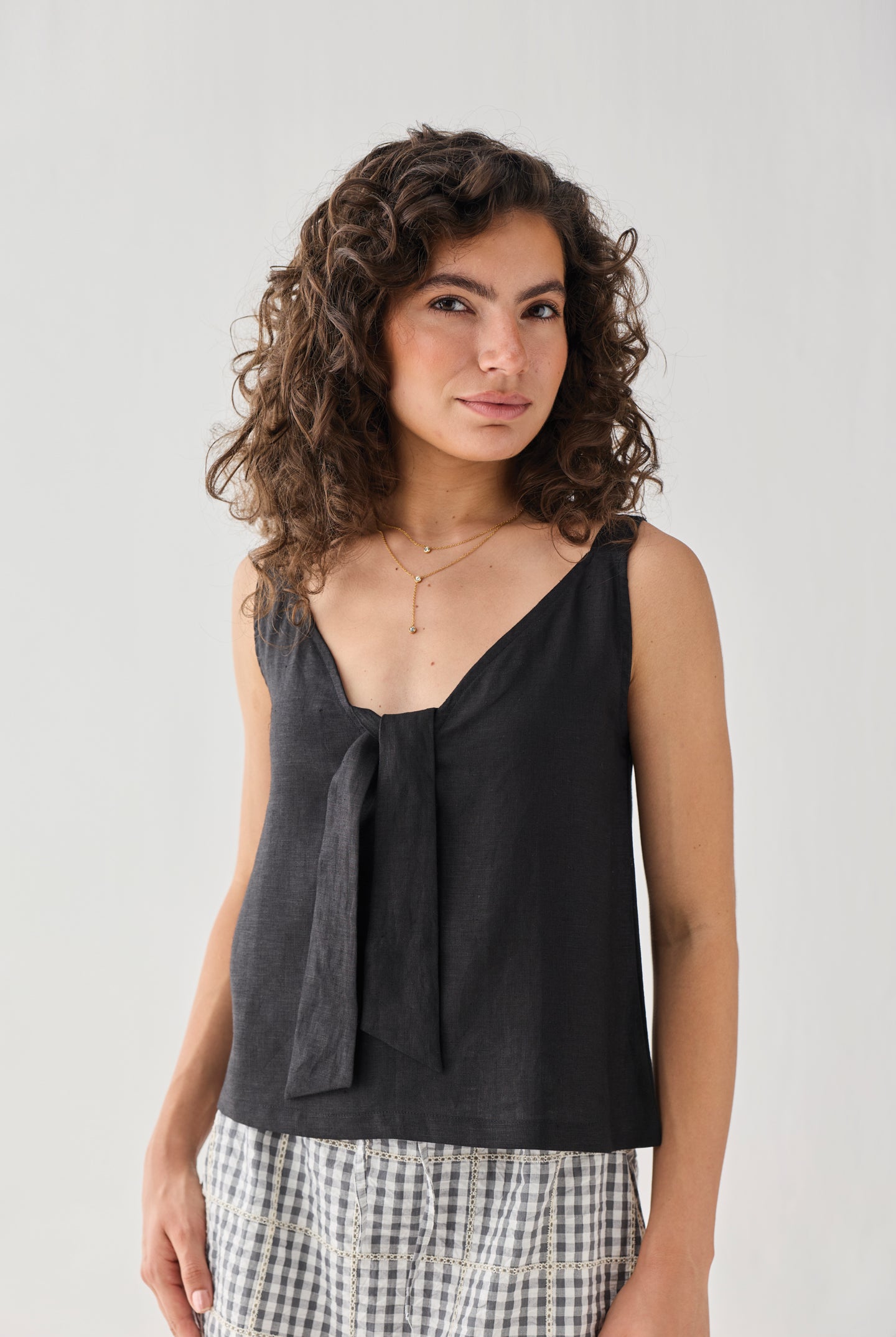 Hemp Front Tie Cami Top in Black with a relaxed, sleeveless silhouette and elegant front tie detail at the V-neckline. Crafted from breathable hemp fabric, the top is shown from the front and paired with a check-pattern skirt.