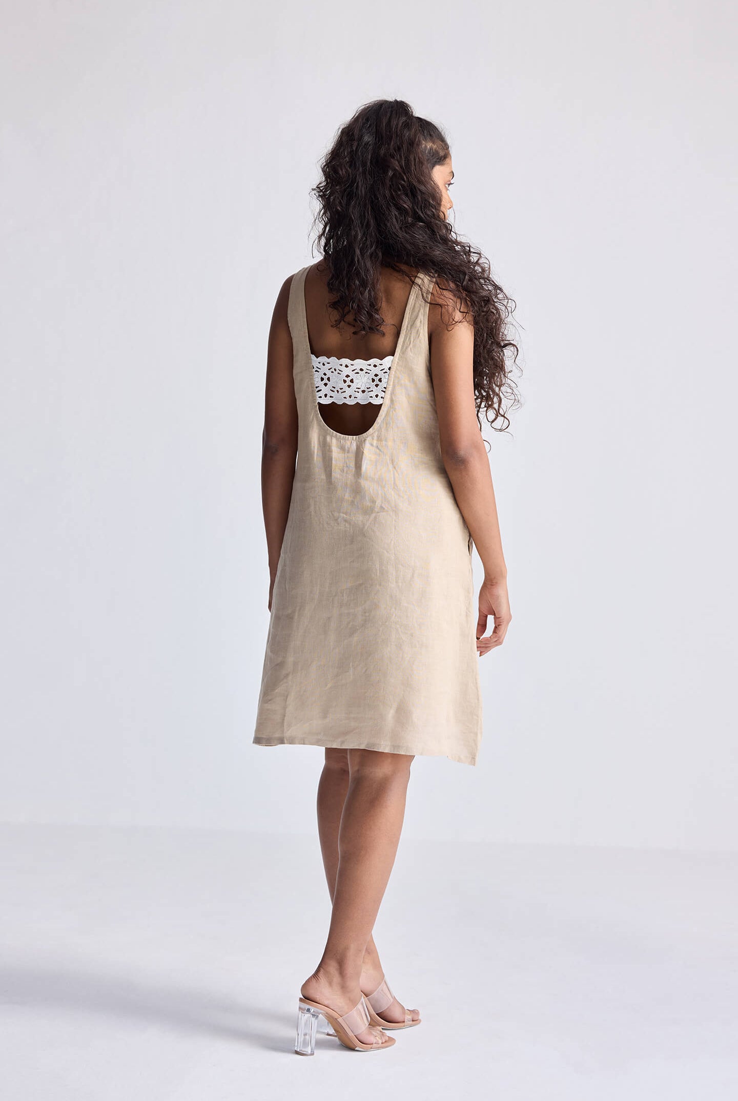 Model wearing the reistor Hemp Short Dress with Back Embroidered Lace Detail, featuring a natural beige hemp fabric, sleeveless silhouette, open back design, and delicate embroidered lace, styled with minimal heels.