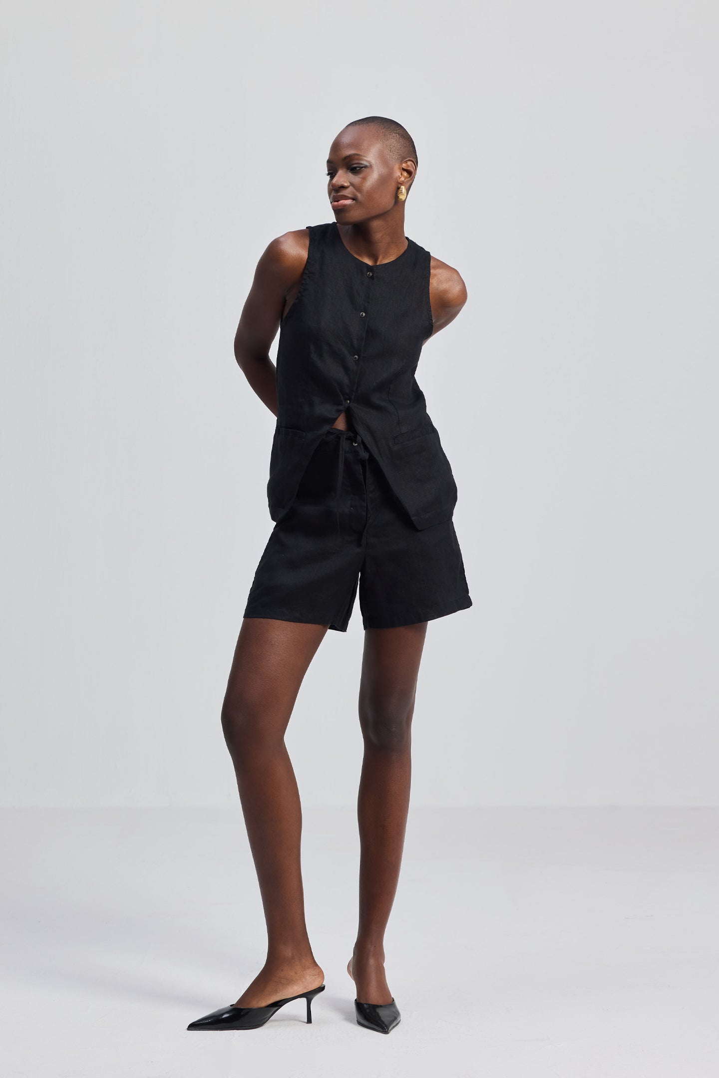 A full-body studio shot of a model wearing the reistor Hemp Vest Jacket Set in Black, featuring a tailored button-up sleeveless vest and matching high-waisted shorts, styled with black pointed-toe kitten heel mules.