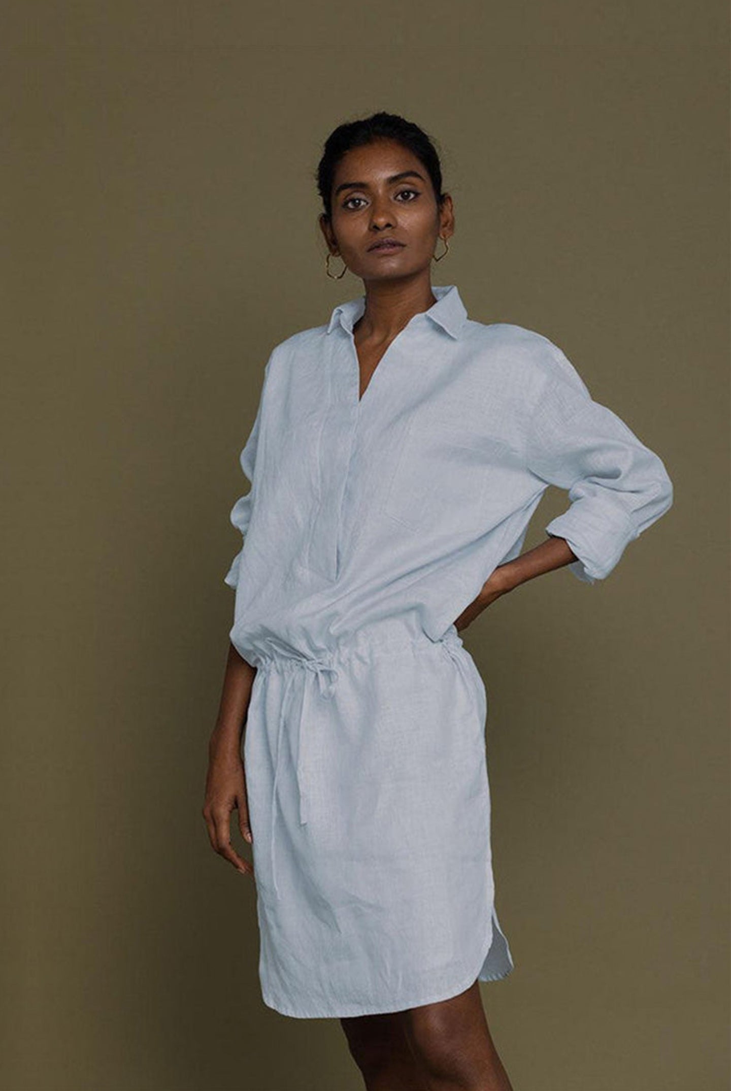 A woman standing against a plain background, wearing a blue collared shirt dress with a front pocket and drawstring waist.