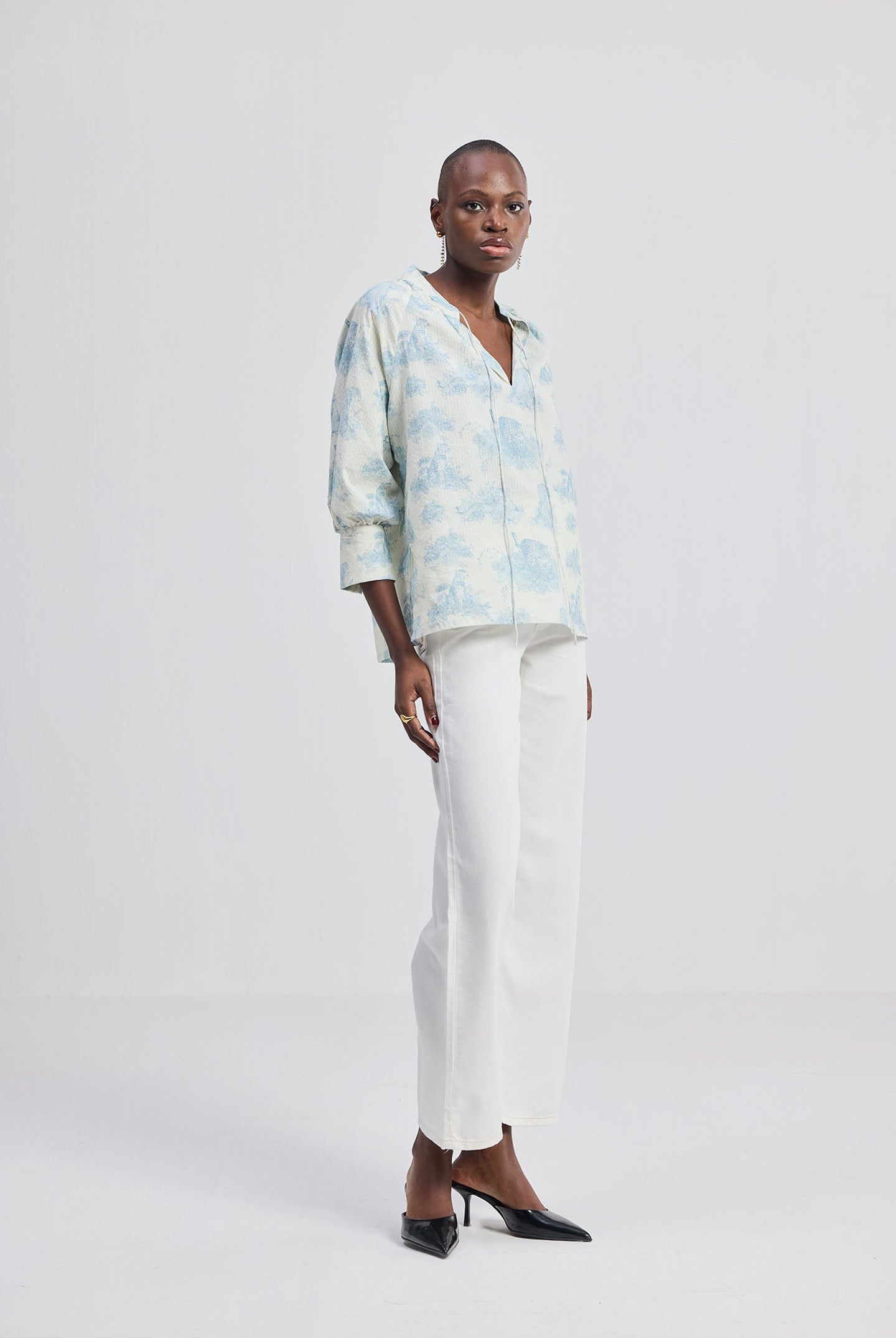 Side-angle full-length shot of a woman wearing the reistor Relaxed-fit Shirt with Drawstring in Animal Print, paired with white trousers and black heels on a minimalist studio background.