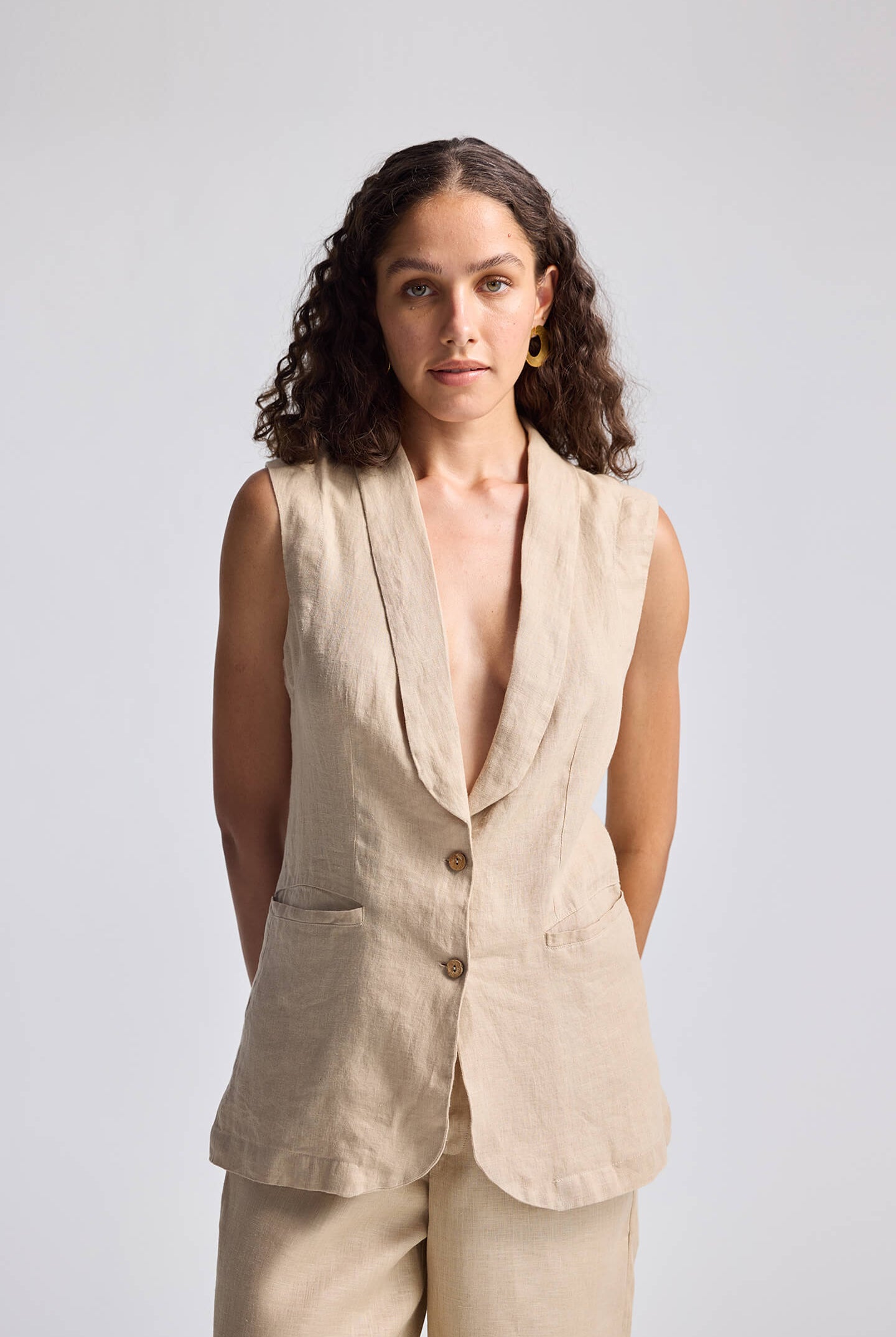 A woman wearing a beige sleeveless hemp jacket with a Shawl Lapel Collar, over a white top, paired with beige trousers.