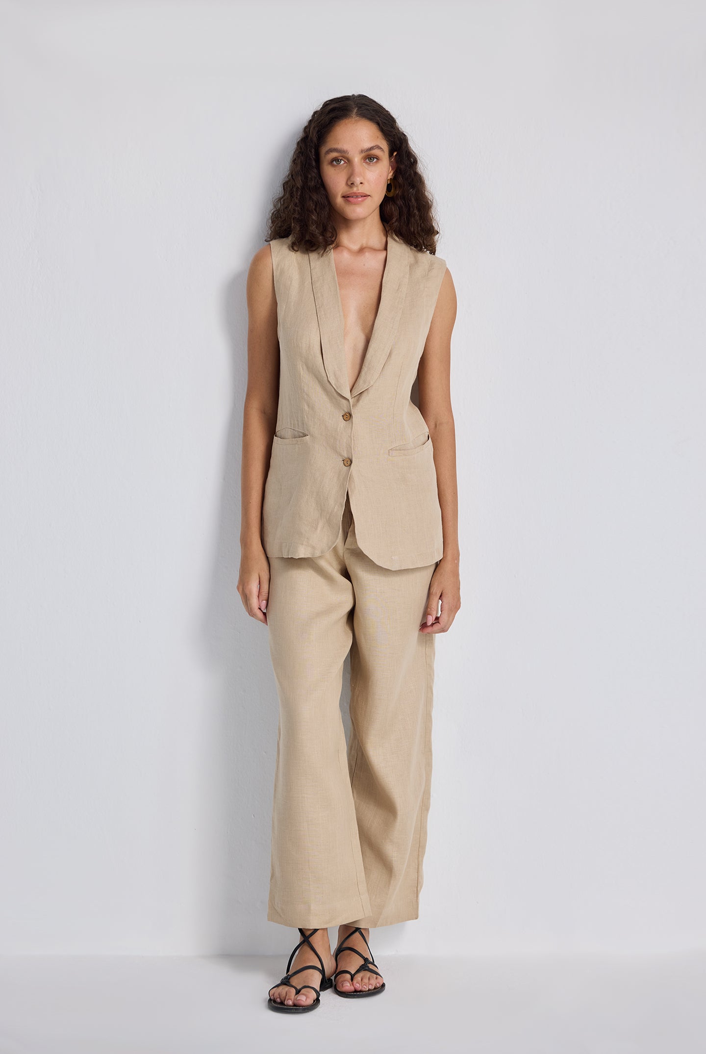 Front view of a model with hands on her hips wearing the beige Sleeveless Hemp Jacket Set by reistor, featuring a button-up longline vest and matching wide-leg trousers.