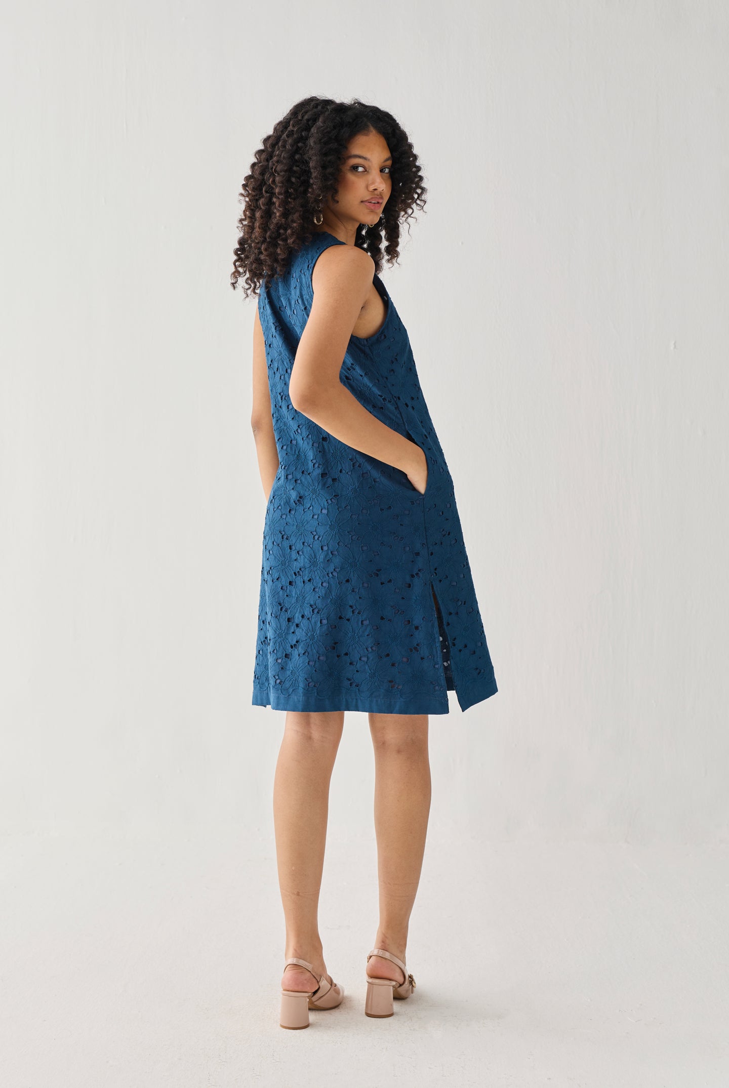Sleeveless Knee-Length Dress in Blue featuring an all-over embroidered eyelet pattern, relaxed A-line silhouette, and practical side pockets, shown from the back in a full-length studio portrait.