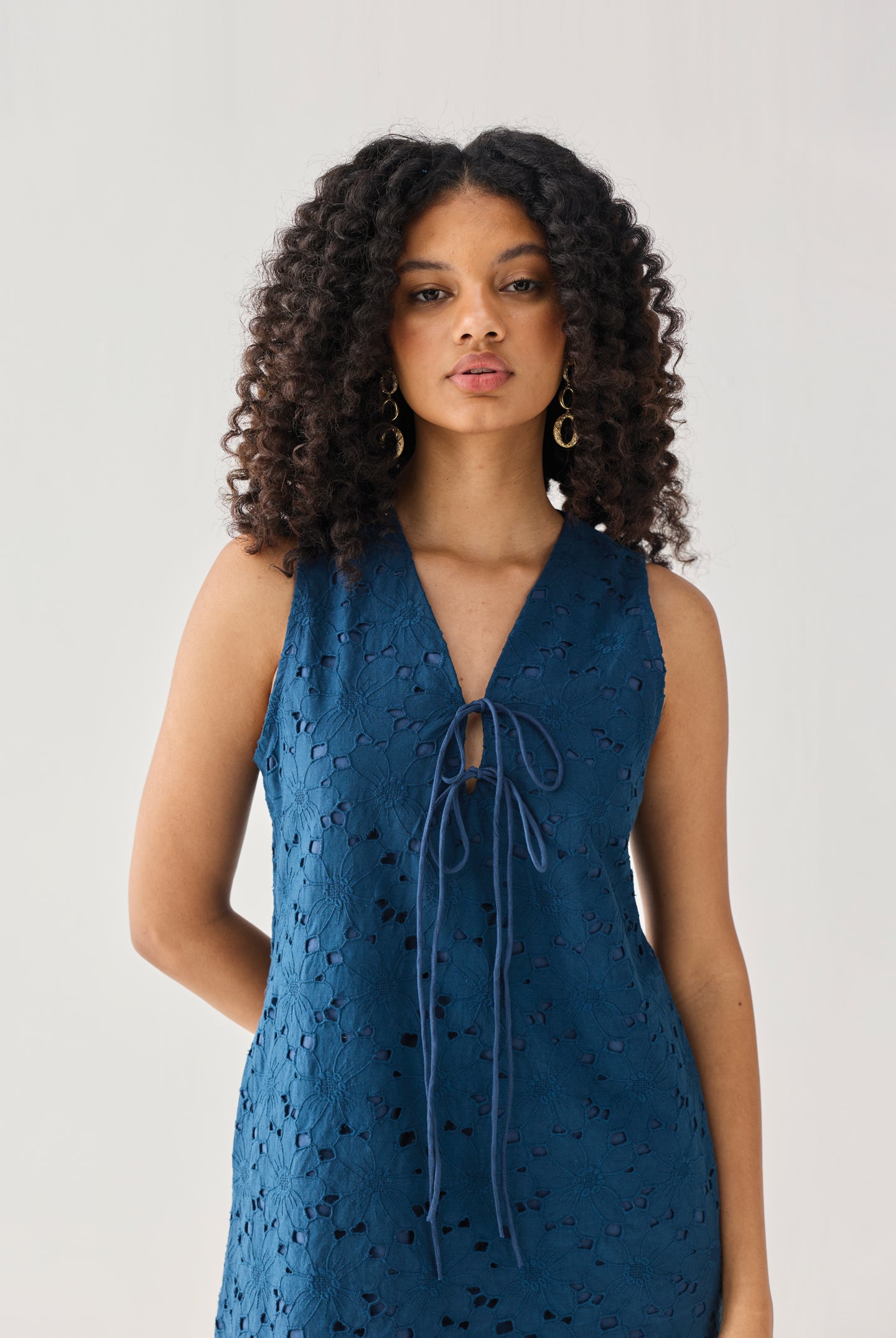 Sleeveless Knee-Length Dress in Blue featuring intricate all-over eyelet embroidery, a V-neckline with delicate self-tie detailing, and a flattering sleeveless silhouette, shown from the front in a studio portrait highlighting the textured fabric and elegant design.