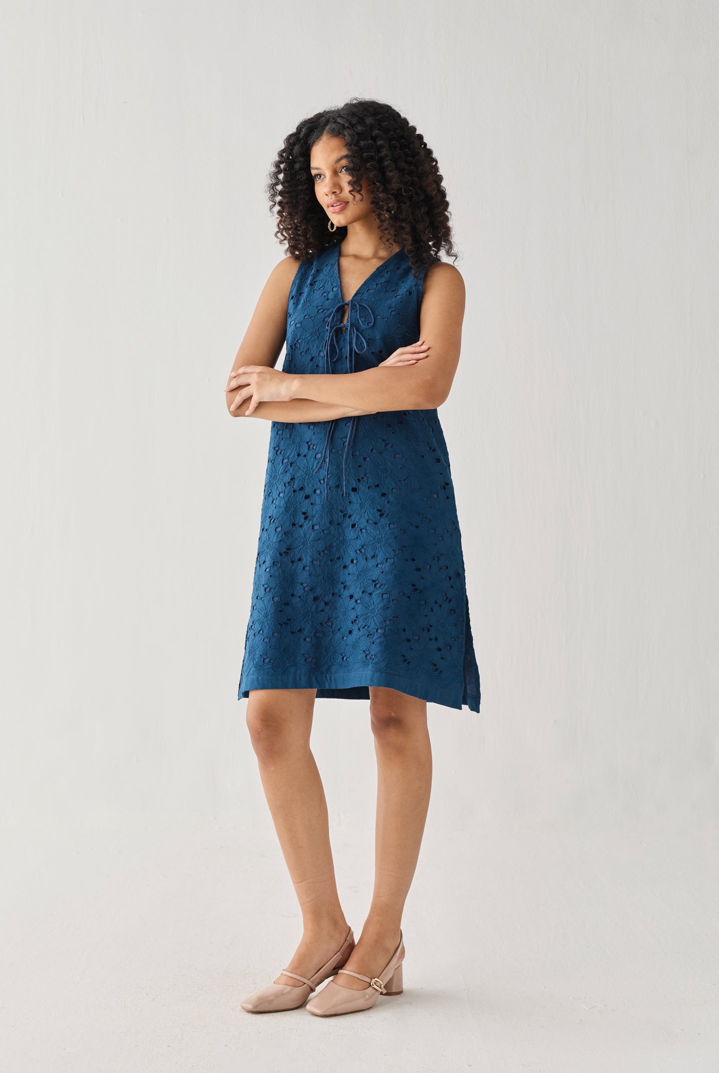 Sleeveless Knee-Length Dress in Blue featuring intricate all-over eyelet embroidery, a V-neckline with self-tie detail, and a relaxed A-line silhouette, shown in a full-length studio portrait that highlights its effortless fit and elegant texture.