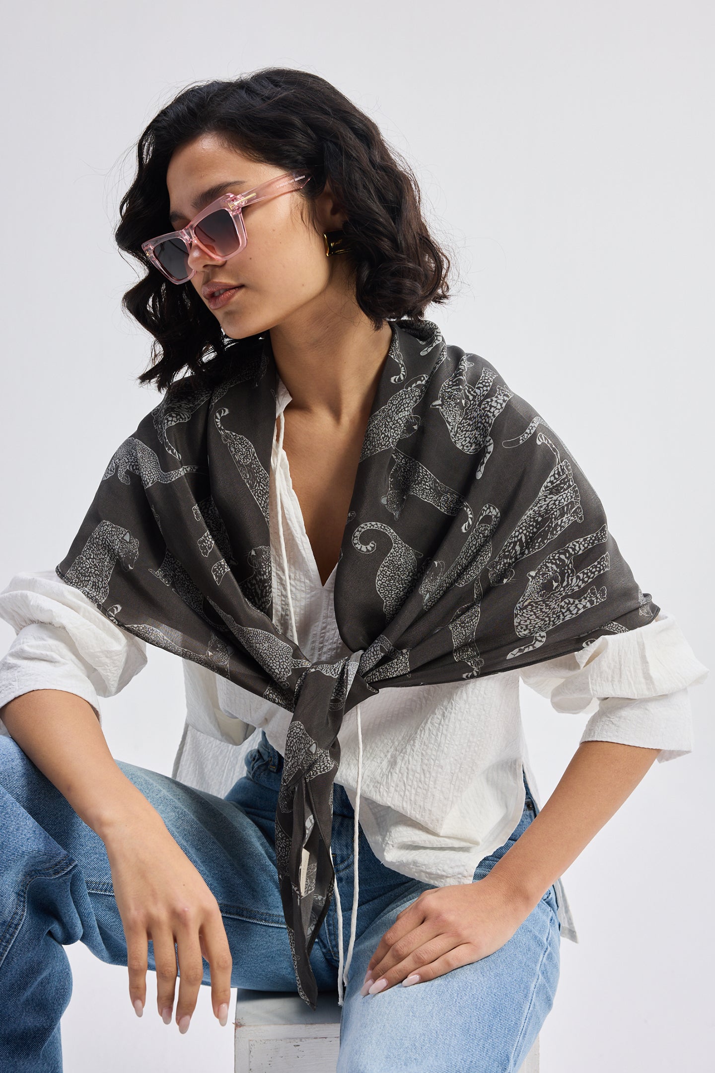 The Mara Scarf in Black styled over a white shirt and blue jeans, worn by a model with pink sunglasses in a casual seated pose.