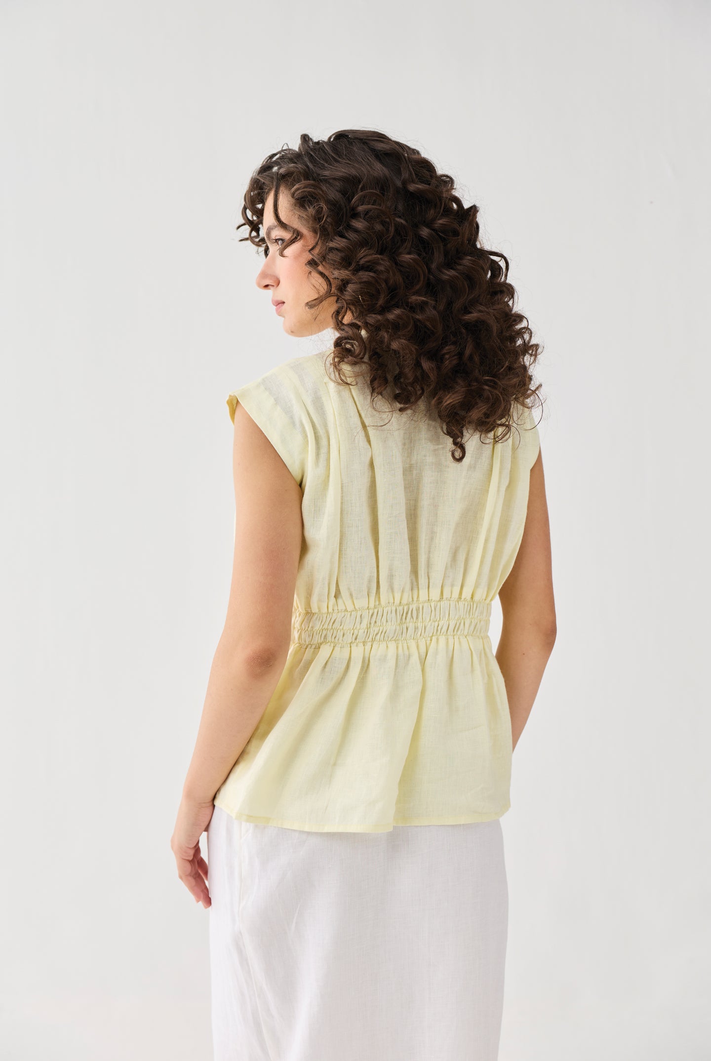 Back view of Reistor's V-neck Overlap Top in Butter Lemon, featuring a smocked waist detail and cap sleeves, styled with a white skirt.