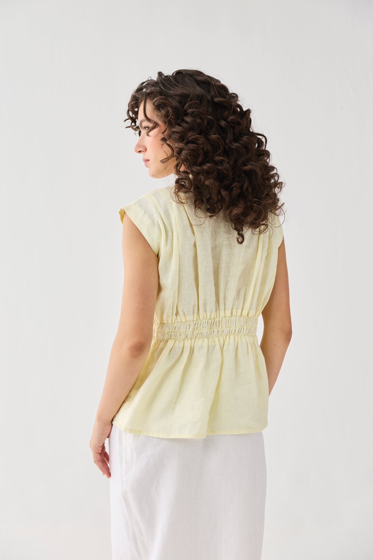 Back view of Reistor's V-neck Overlap Top in Butter Lemon, featuring a smocked waist detail and cap sleeves, styled with a white skirt.