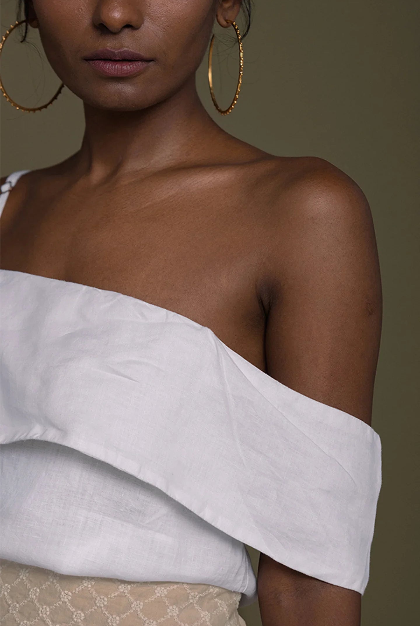 Woman wearing a Reistor white off-shoulder top with gold hoop earrings on a brown background.