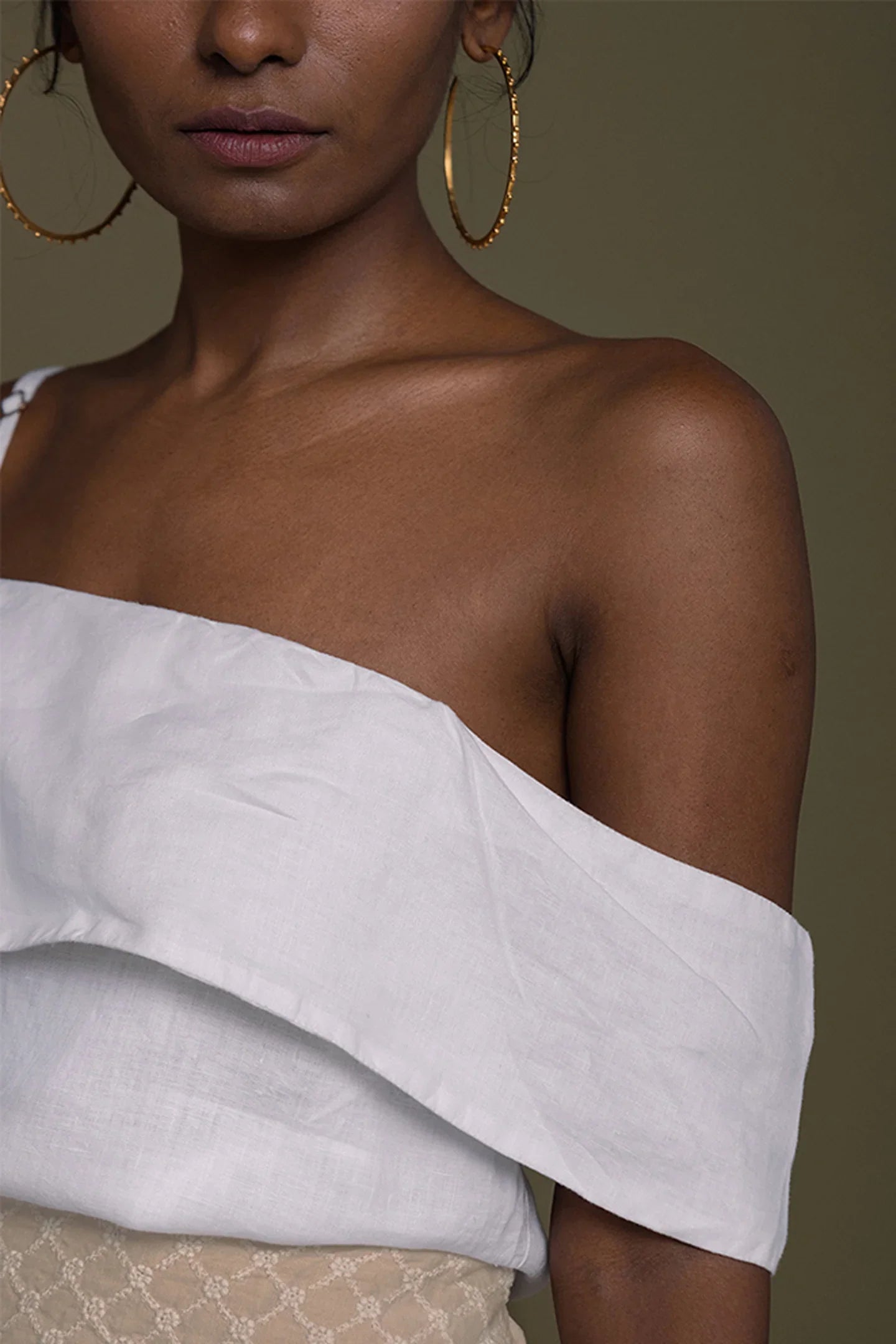 Woman wearing a Reistor white off-shoulder top with gold hoop earrings on a brown background.