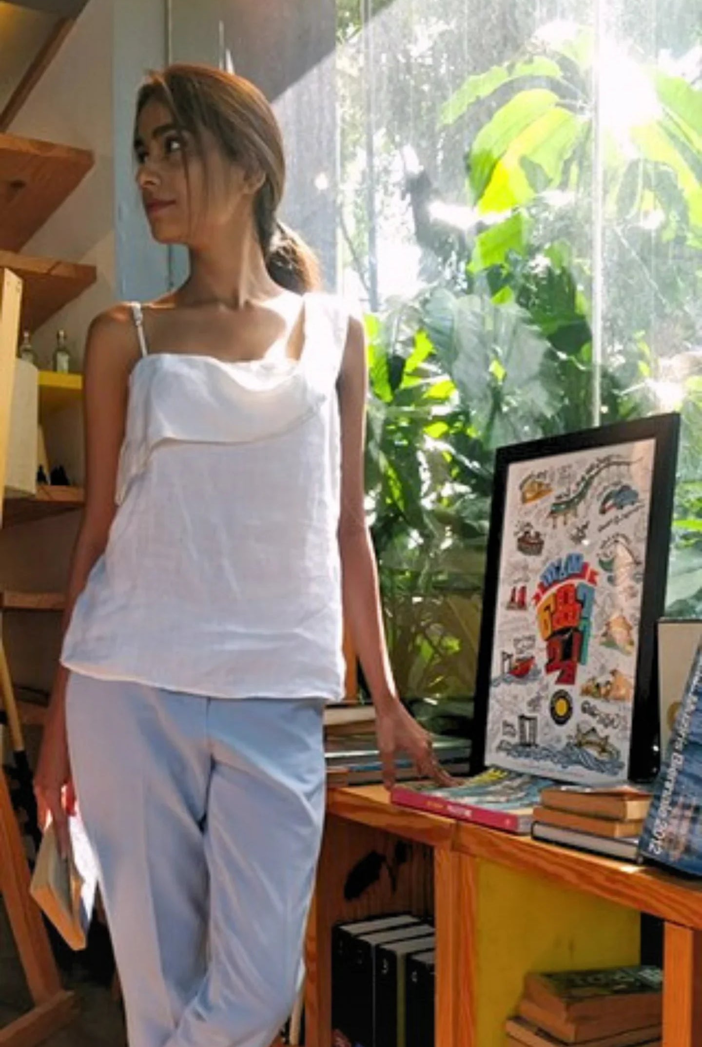 Woman wearing a Reistor white top and light blue pants, standing in a room with books and a framed picture.