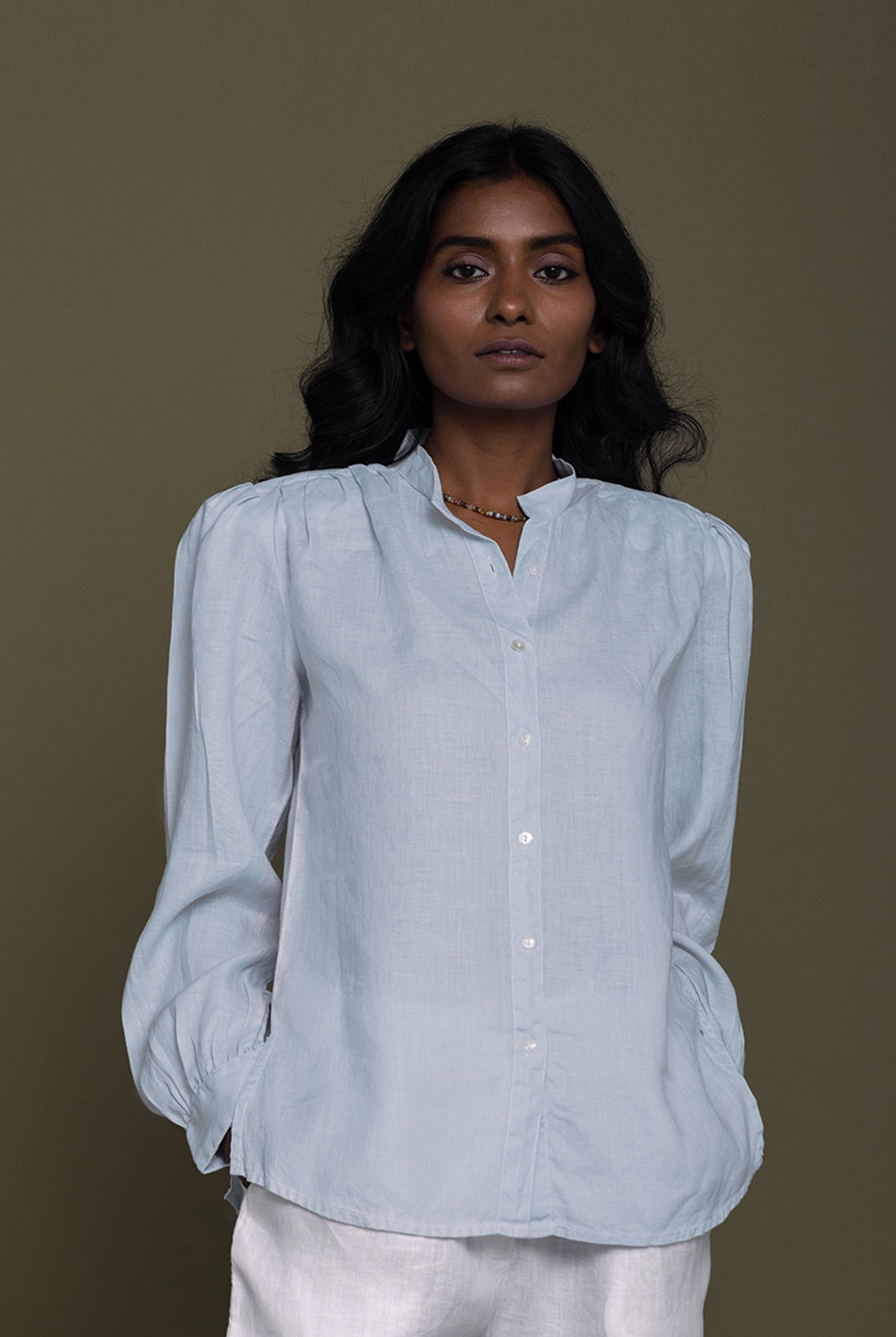 A person standing against a neutral background, wearing a blue pleated button-down shirt with shoulder pads and cuff gathers, and a necklace.