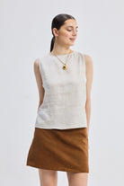 A woman wearing a beige boat neck top with side cut-outs, paired with a brown skirt. She is accessorized with a necklace and is standing against a white background.