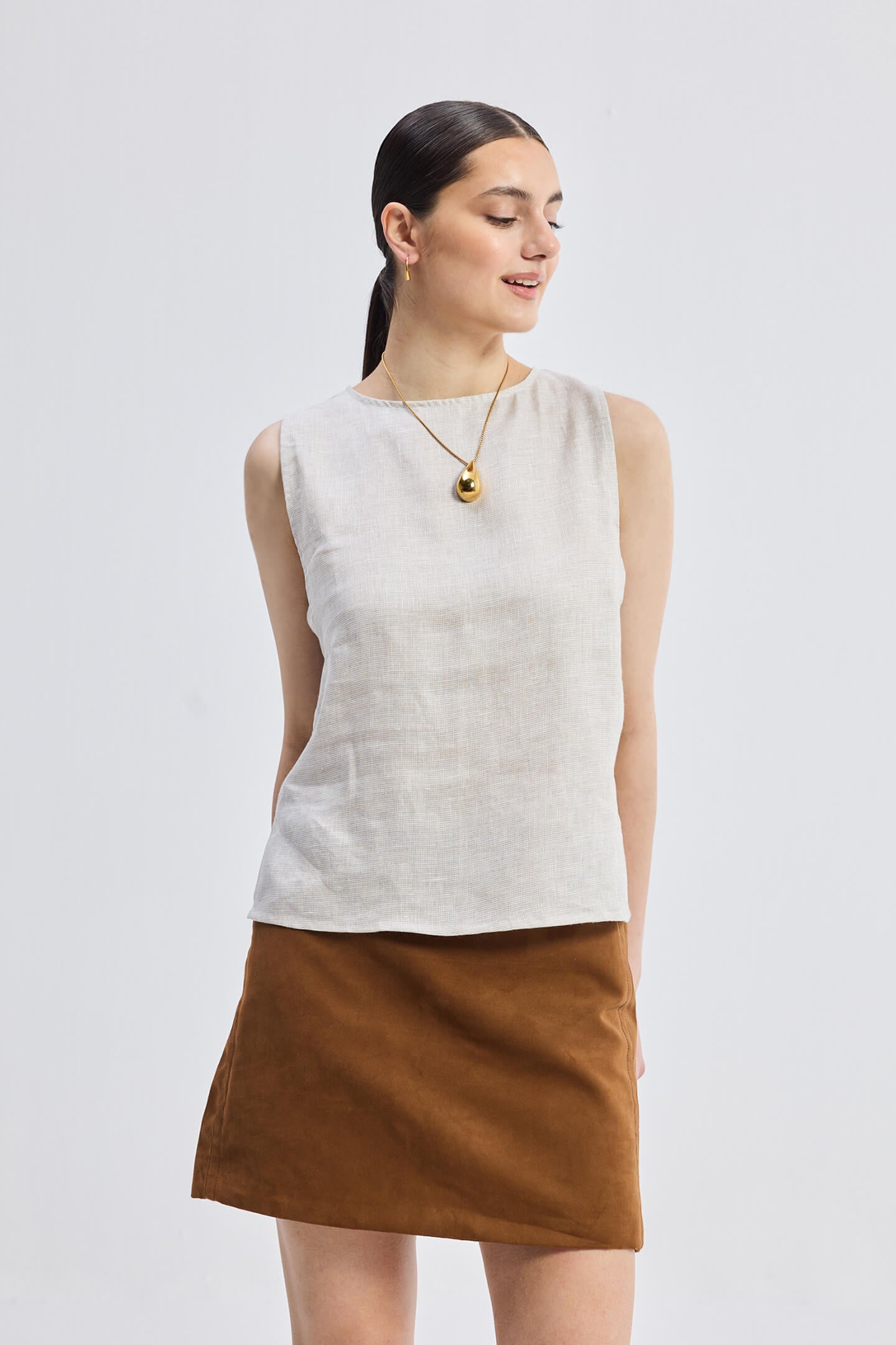 A woman wearing a beige boat neck top with side cut-outs, paired with a brown skirt. She is accessorized with a necklace and is standing against a white background.