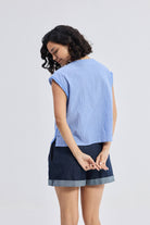 Model wearing Reistor Boxy V-neck Top with Fray Detail in blue textured fabric, relaxed fit sleeveless sustainable women’s top paired with dark cuffed shorts, back view.