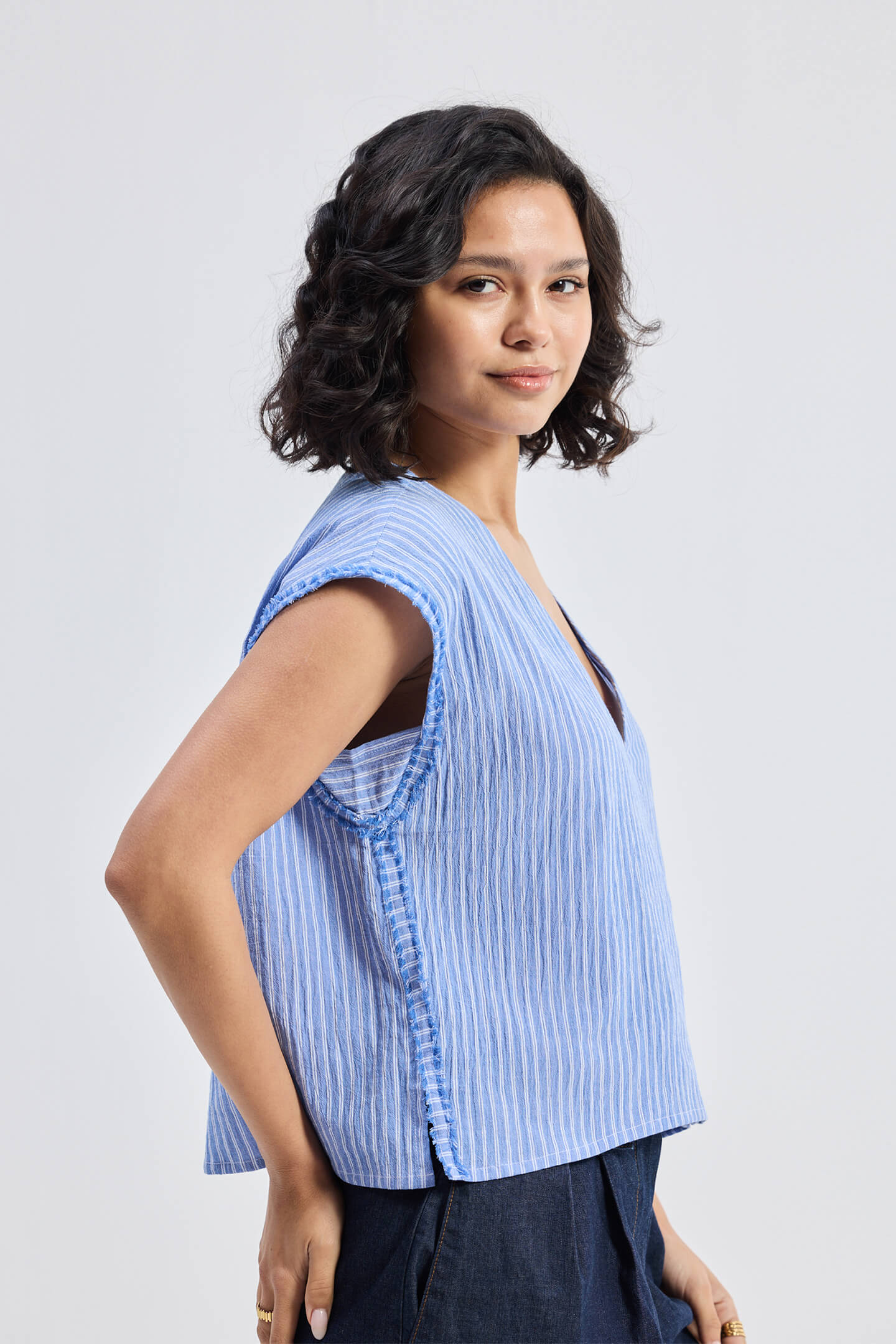 Model wearing Reistor Boxy V-neck Top with Fray Detail in blue textured fabric, relaxed fit sleeveless sustainable women’s top, side view.