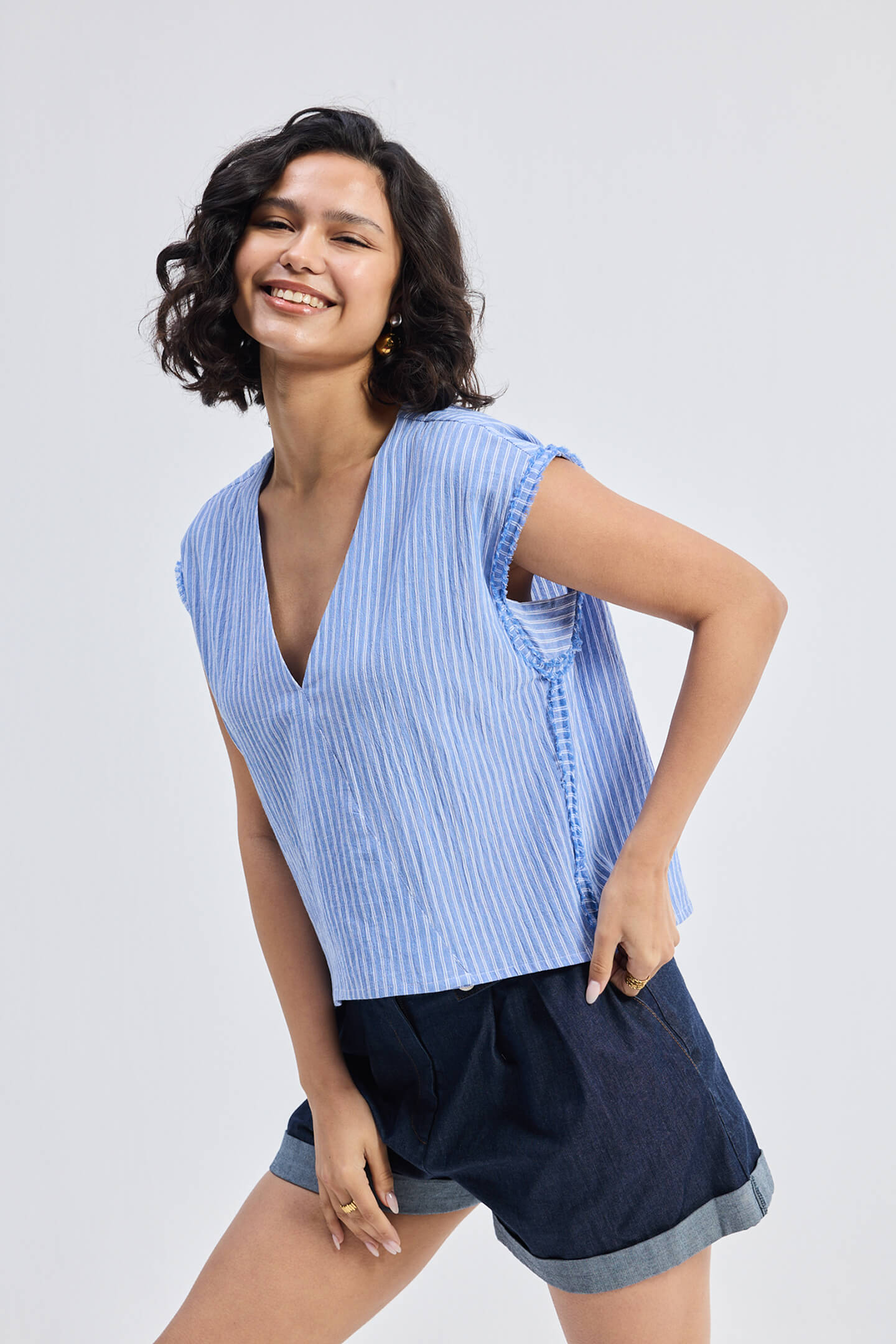 Smiling model wearing Reistor Boxy V-neck Top with Fray Detail in blue textured fabric, relaxed fit sleeveless eco-friendly women’s top styled with dark cuffed shorts.