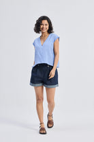 Full-length view of model wearing Reistor Boxy V-neck Top with Fray Detail in blue textured fabric, relaxed fit eco-friendly women’s top paired with dark cuffed shorts and sandals.