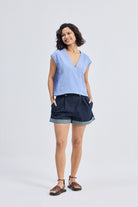 Front full-length view of model wearing Reistor Boxy V-neck Top with Fray Detail in blue striped textured fabric, relaxed fit sustainable women’s top styled with dark cuffed shorts and sandals.
