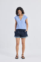 Model wearing Reistor Boxy V-neck Top with Fray Detail in blue striped textured fabric, relaxed fit eco-friendly women’s top paired with dark cuffed shorts and sandals, front view.