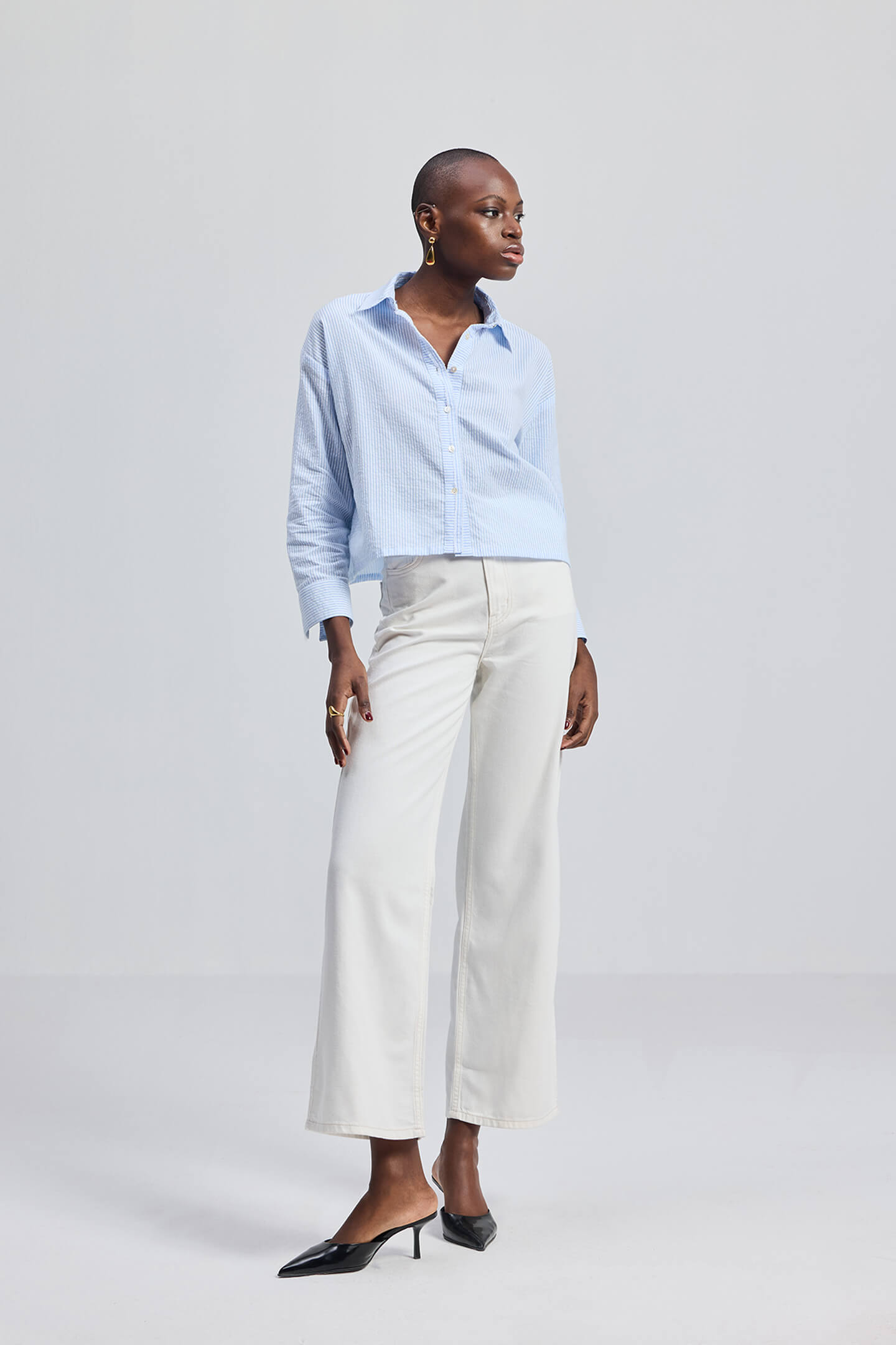 A person wearing a light blue, long-sleeved, button-up shirt and white trousers. The person is standing against a plain background.