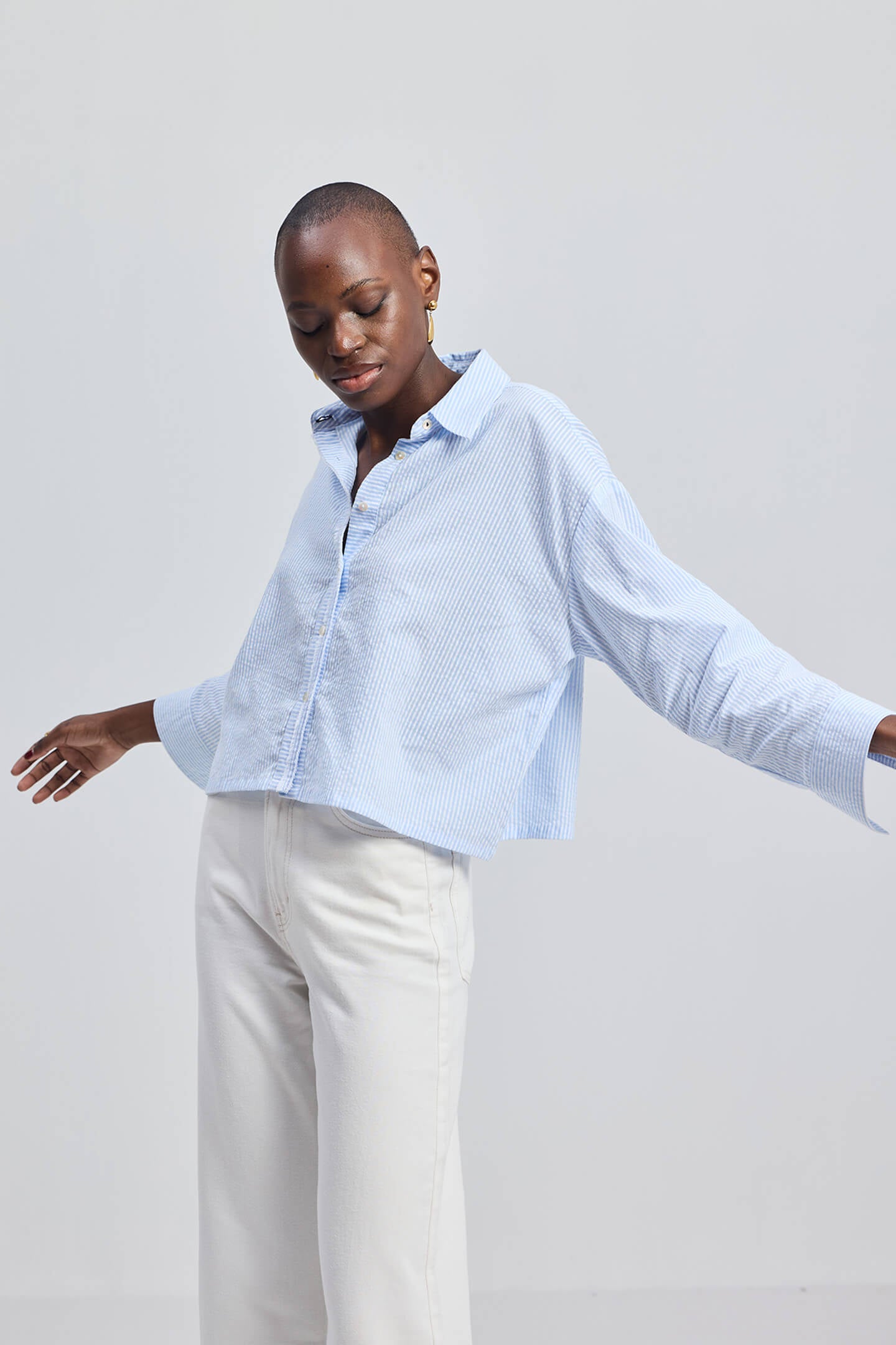 A model wearing the Reistor Crop it like it’s Hot Shirt in Stripes, featuring light blue and white vertical stripes, a button-down front, and a relaxed cropped fit paired with white trousers.